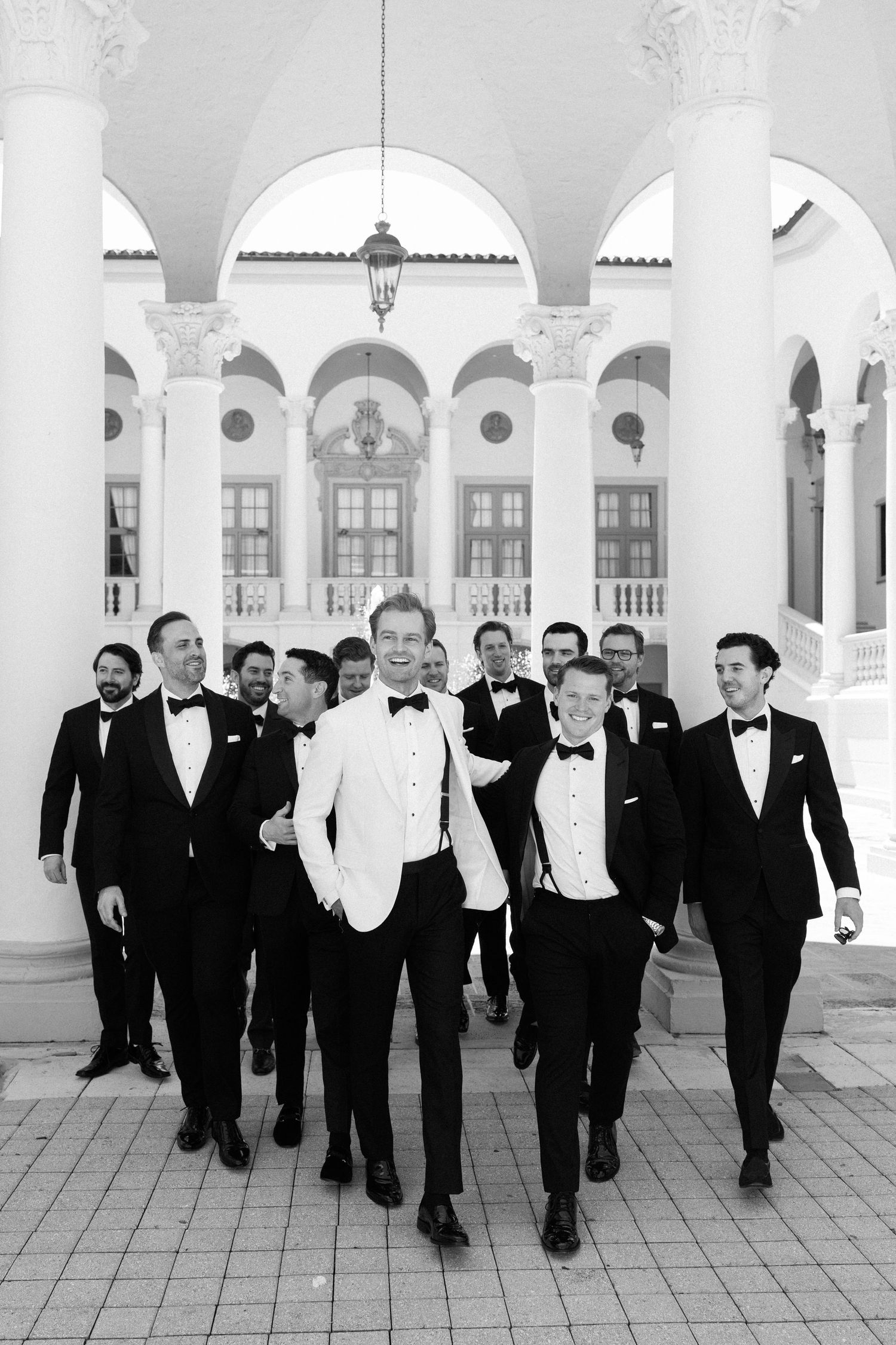 Groom in white tuxedo jacket leads groomsmen in black tuxedos walking confidently before elegant columned venue.