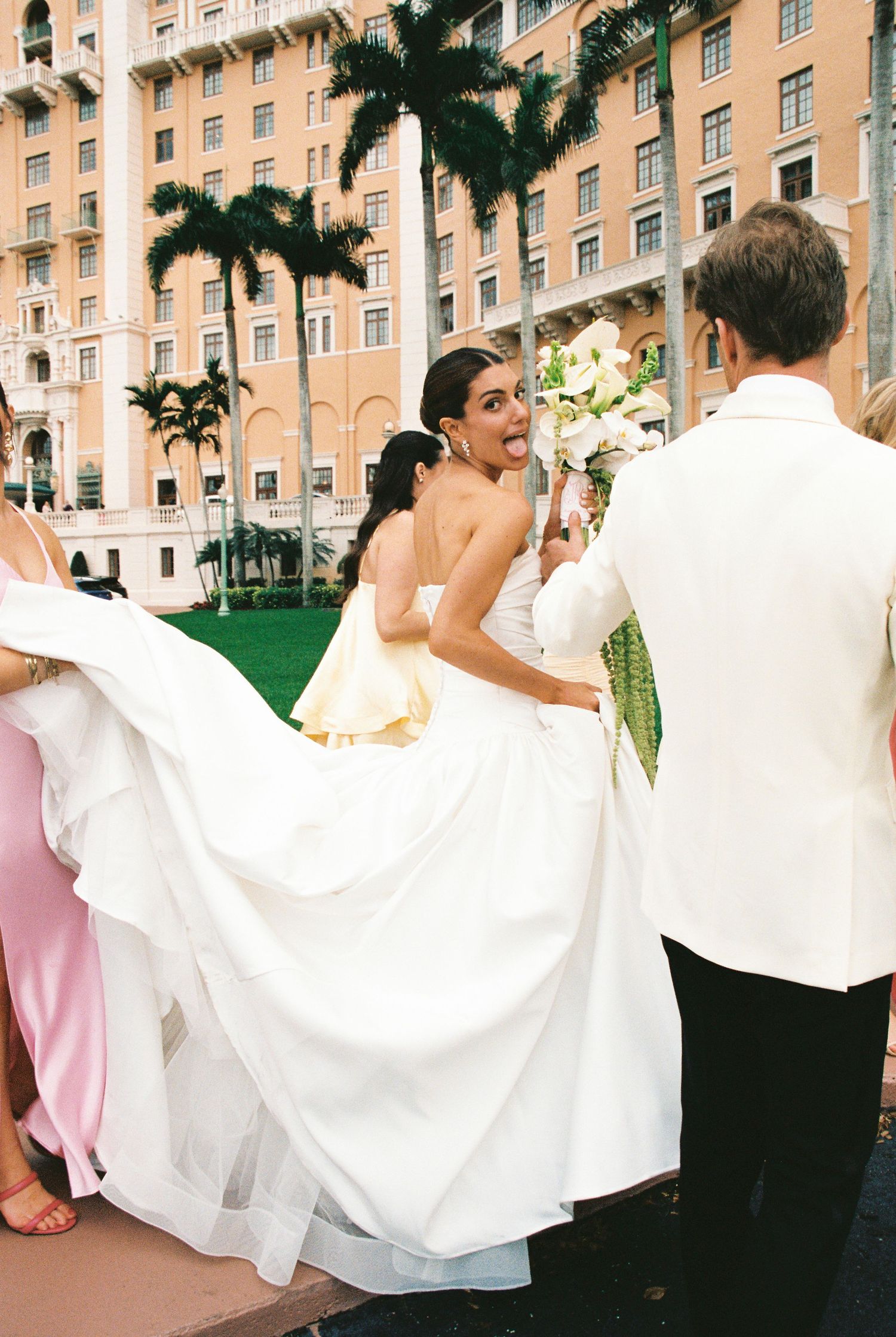 Smiling bride in strapless white gown holds bouquet as bridesmaids lift her dress outside a luxury hotel.