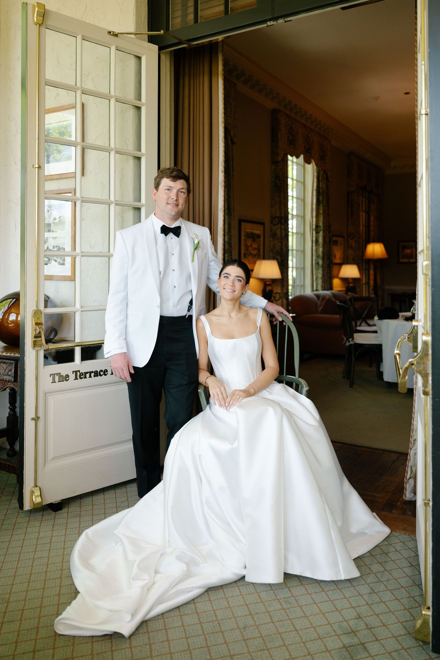 Bride in white ballgown sits beside groom in white tuxedo jacket at elegant wedding venue doorway.