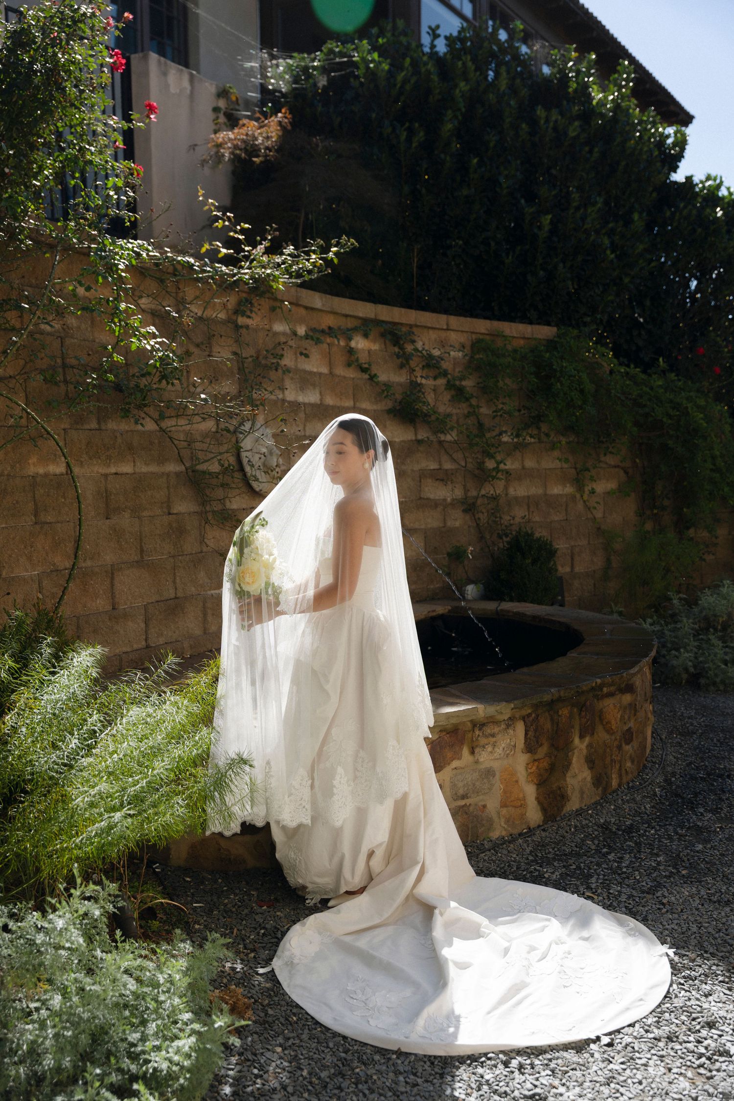 A figure in a flowing white wedding dress and veil poses gracefully in a garden setting against stone walls.