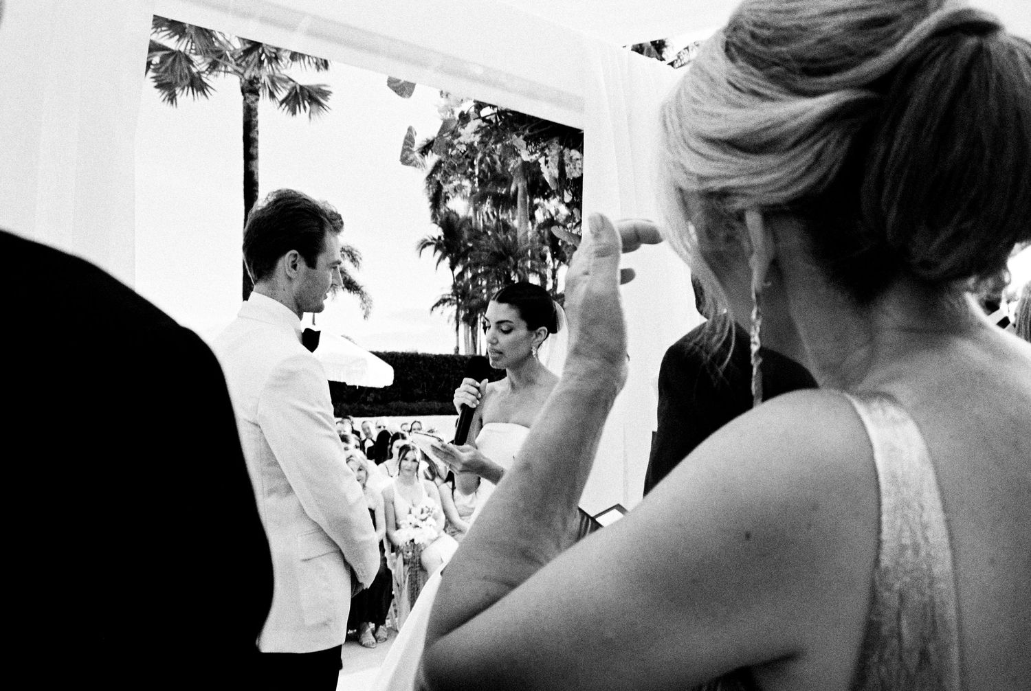 Black and white wedding ceremony photo showing couple exchanging vows while guests watch and photograph the moment.