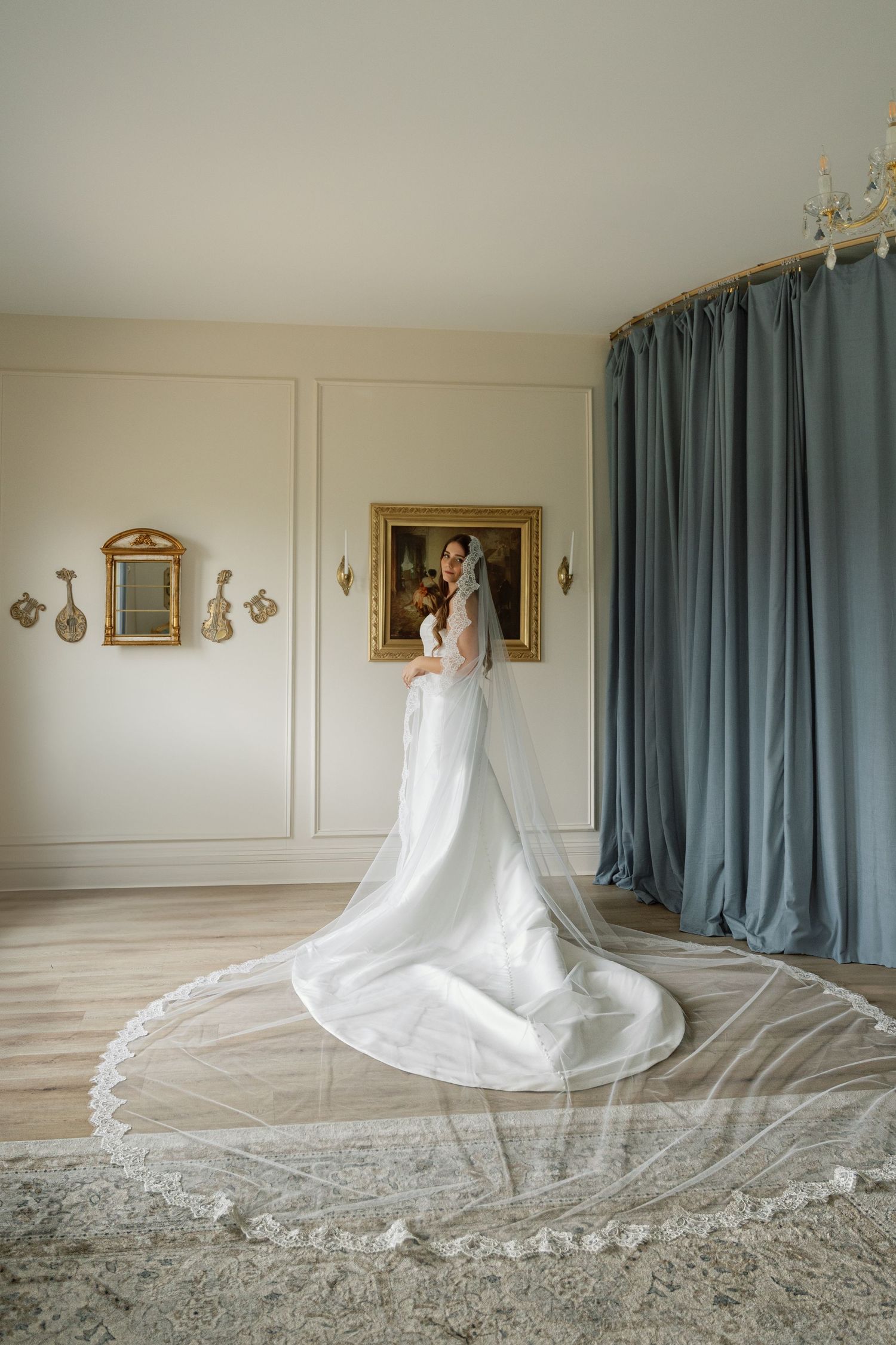 Elegant wedding gown with dramatic cathedral-length lace veil in a room with blue curtains and gold-framed artwork.