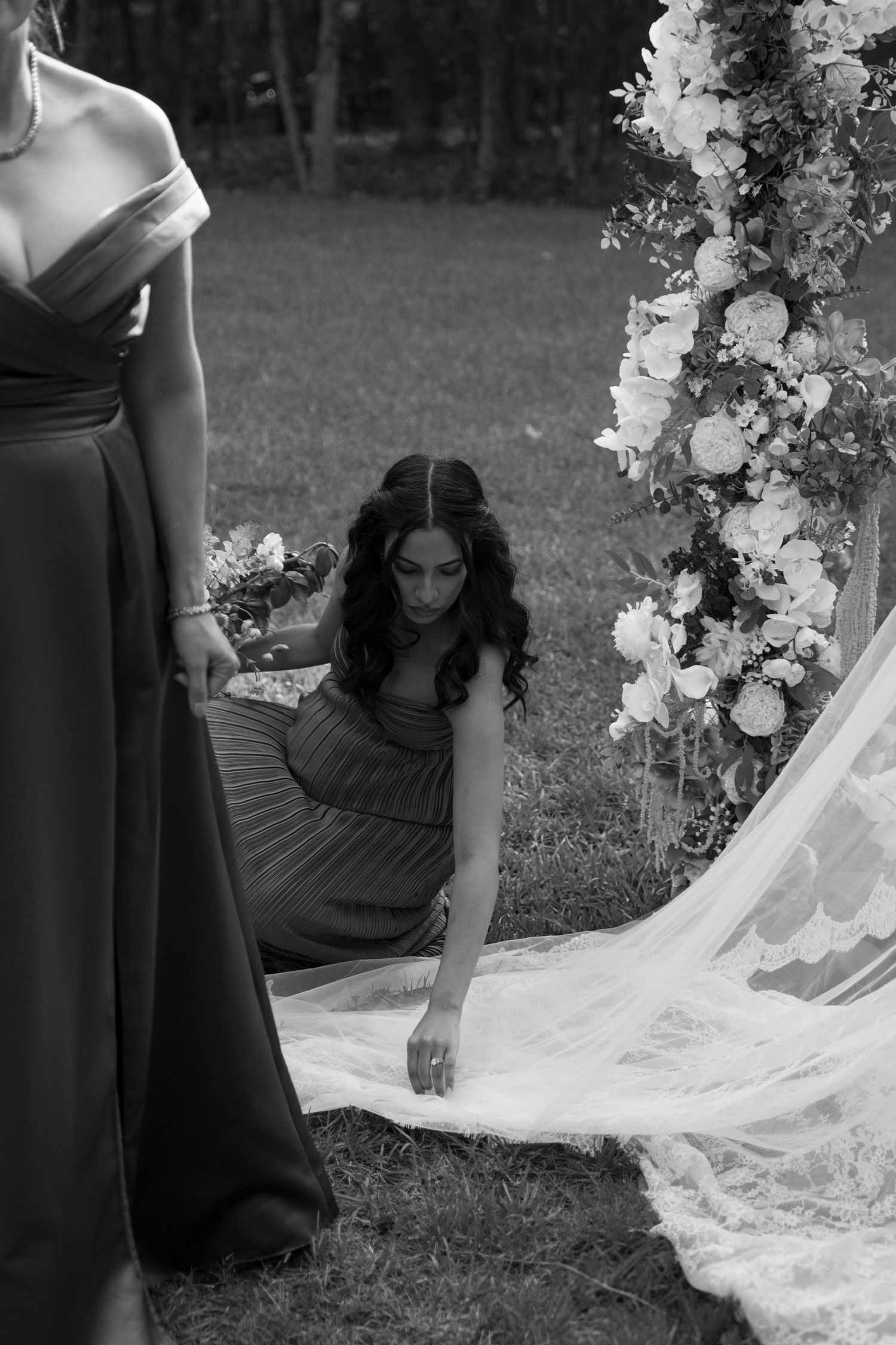 Black and white wedding photo series showing someone arranging a long flowing bridal veil on grass outdoors.