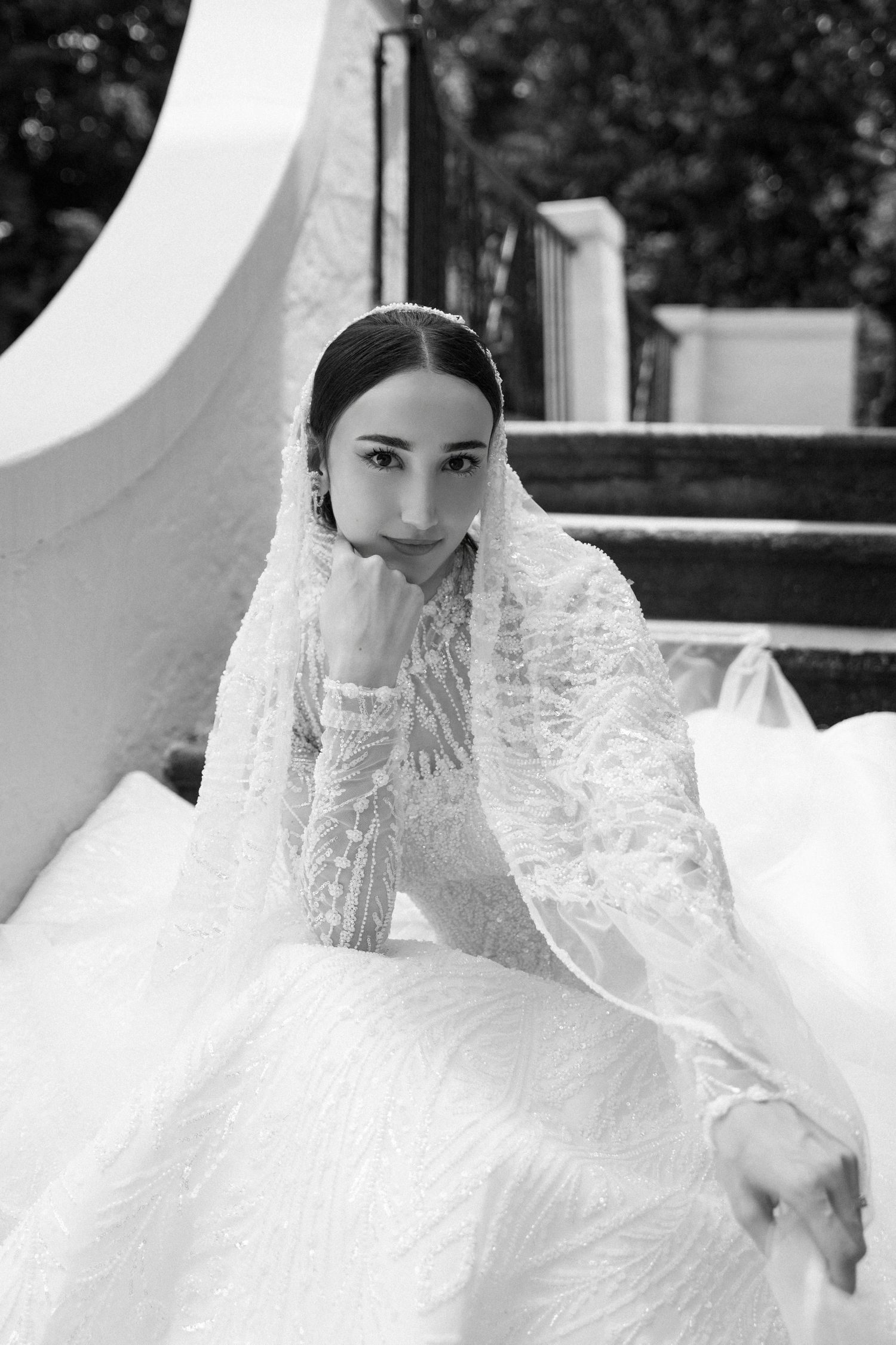 A bride in a flowing white lace wedding dress and veil poses gracefully in an outdoor garden setting.