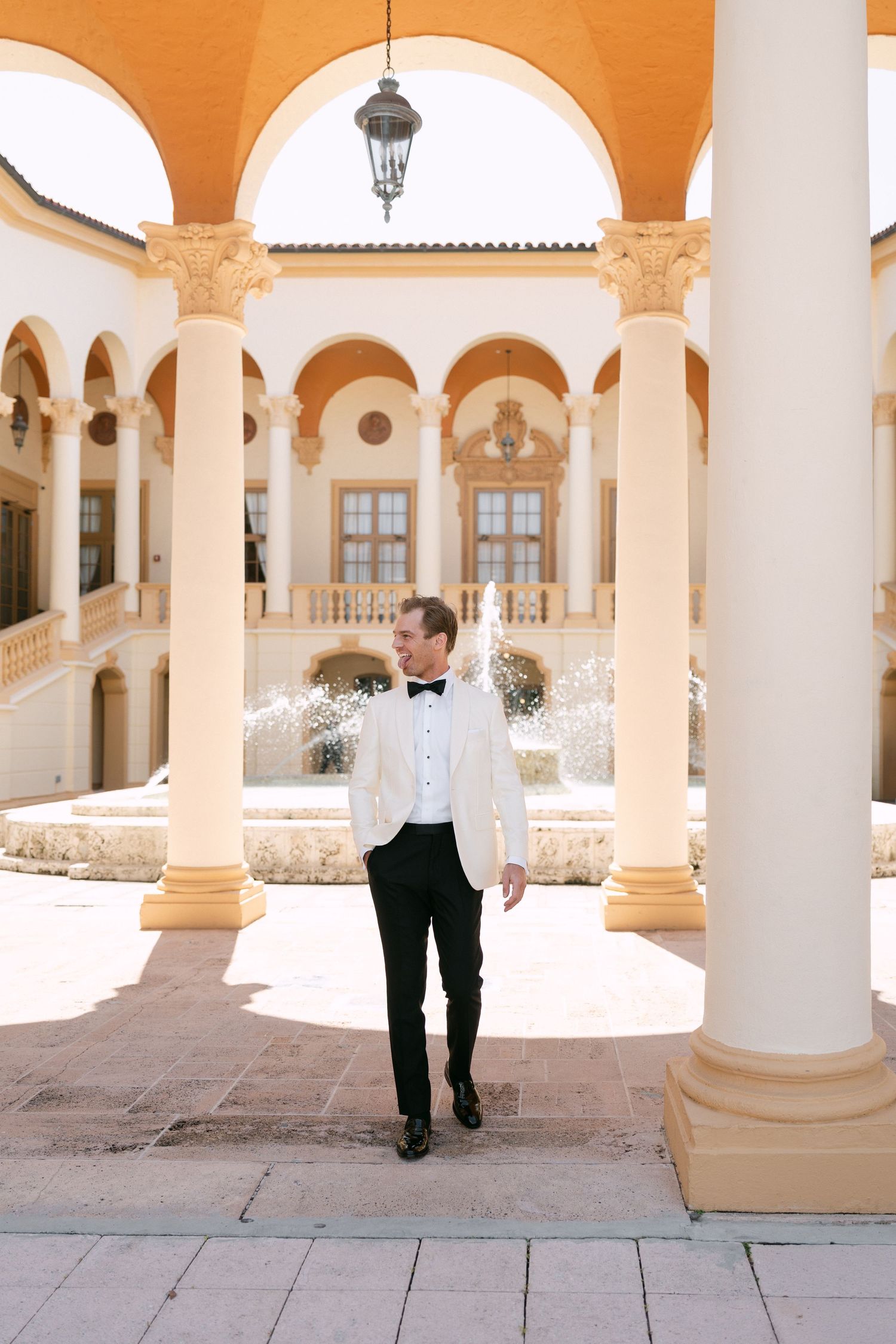 Groom in white tuxedo jacket and black trousers walking in elegant courtyard with arched columns and fountain.
