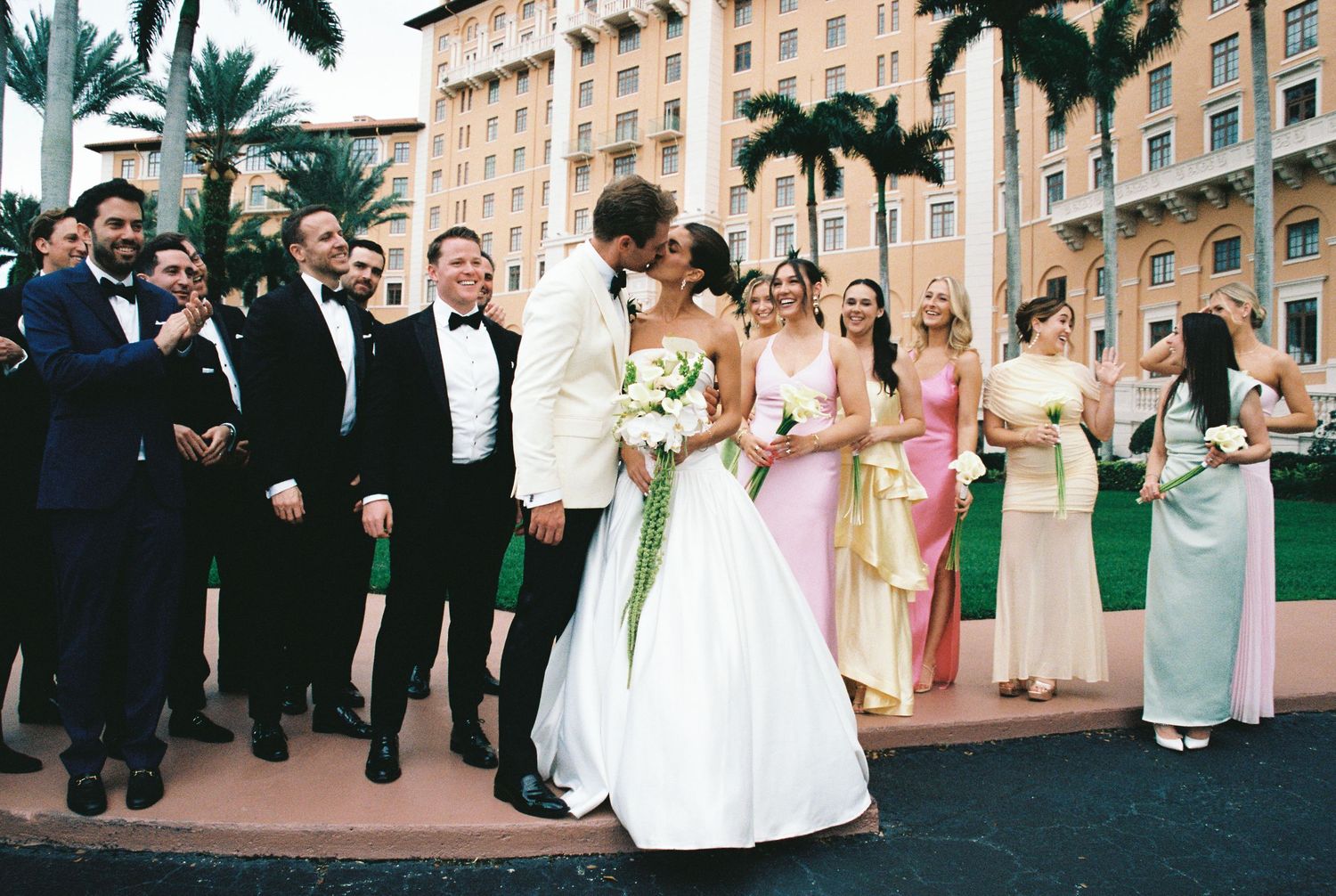 Wedding couple kissing outside Boca Raton Resort, surrounded by bridal party in pink dresses and black tuxedos.