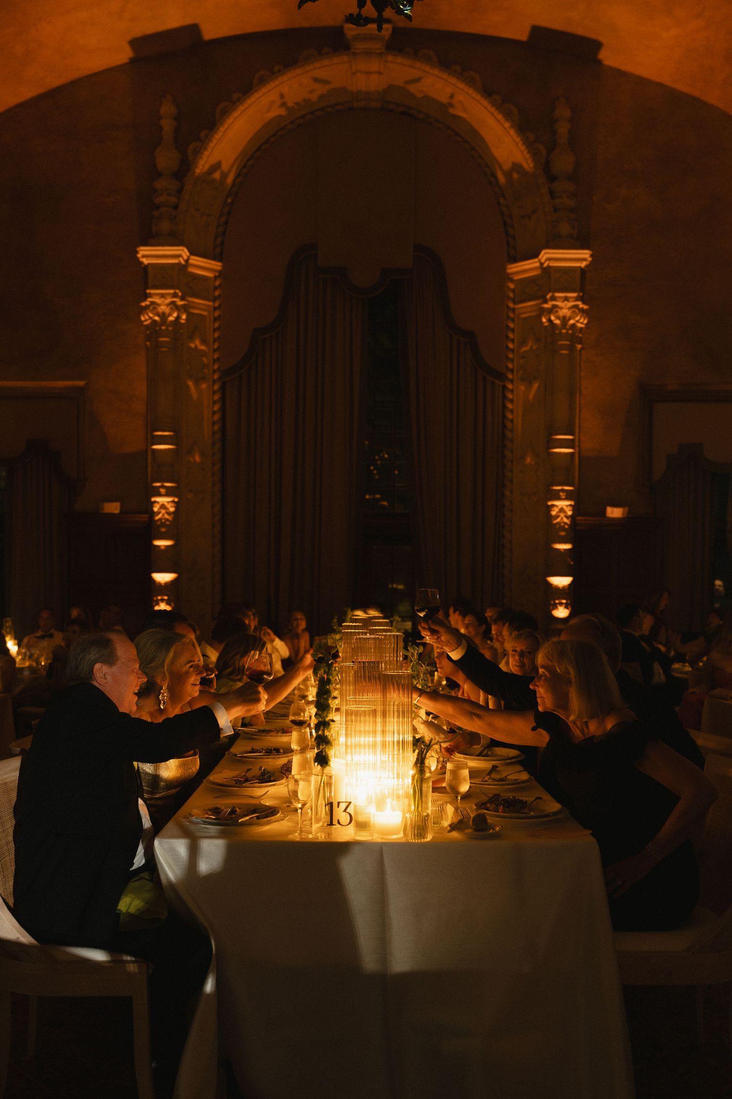 Guests raise glasses in candlelit toast at elegant dinner table inside ornate arched venue with warm golden lighting.