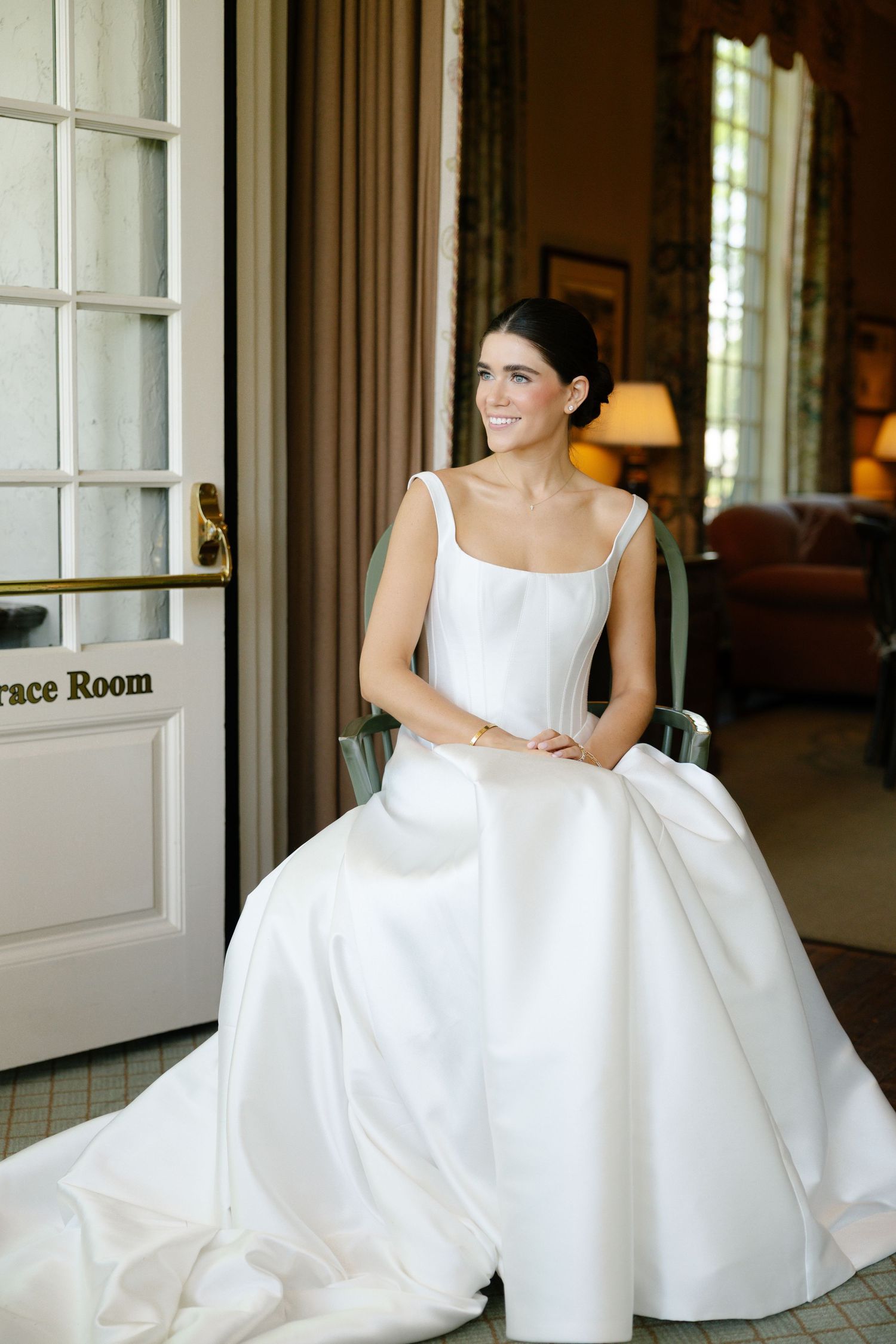 A smiling bride in a elegant white square-neck ballgown sits gracefully in a chair inside a classic wedding venue.