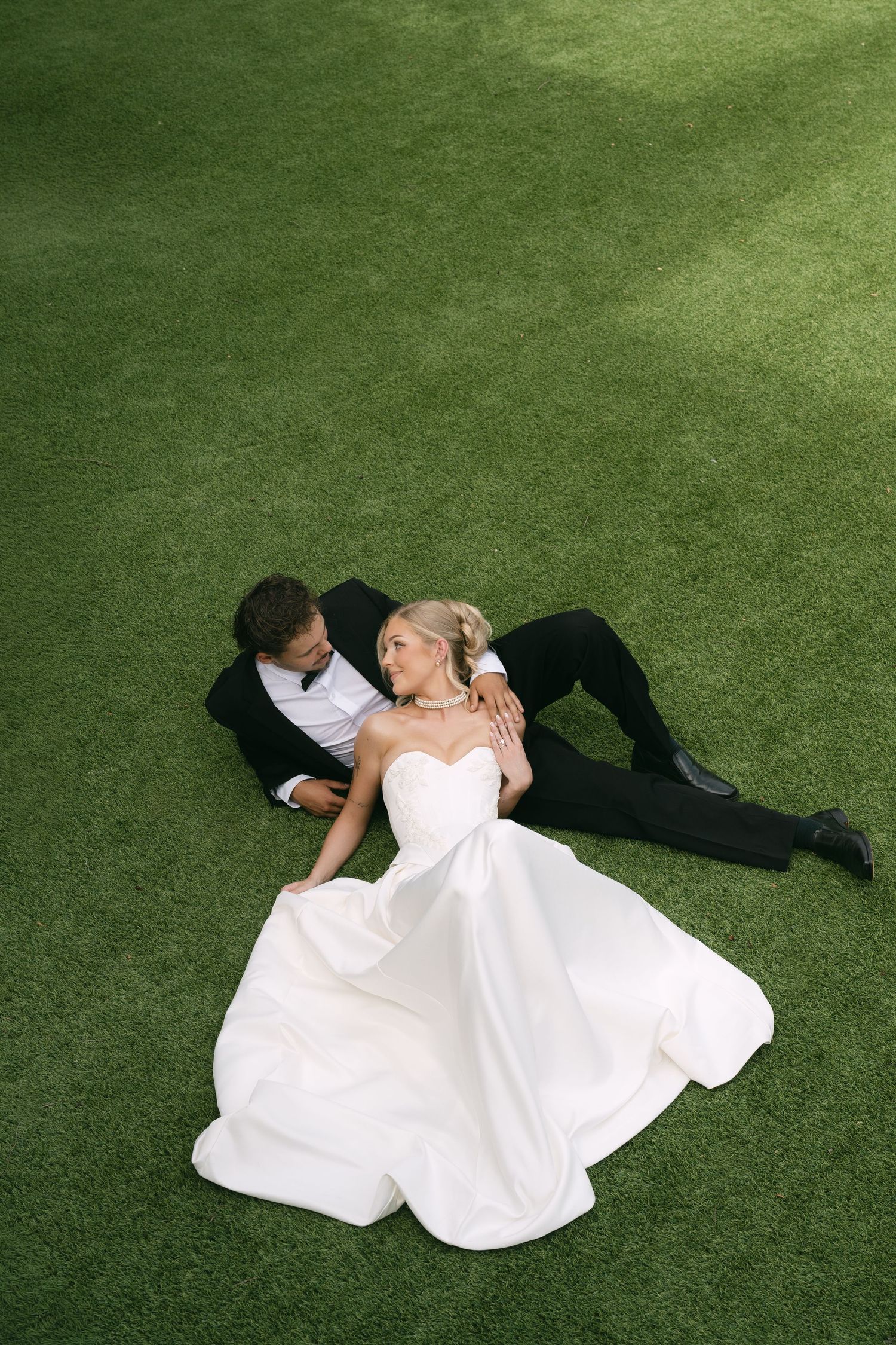 A bride and groom in wedding attire sit together on lush green grass, gazing at each other romantically.