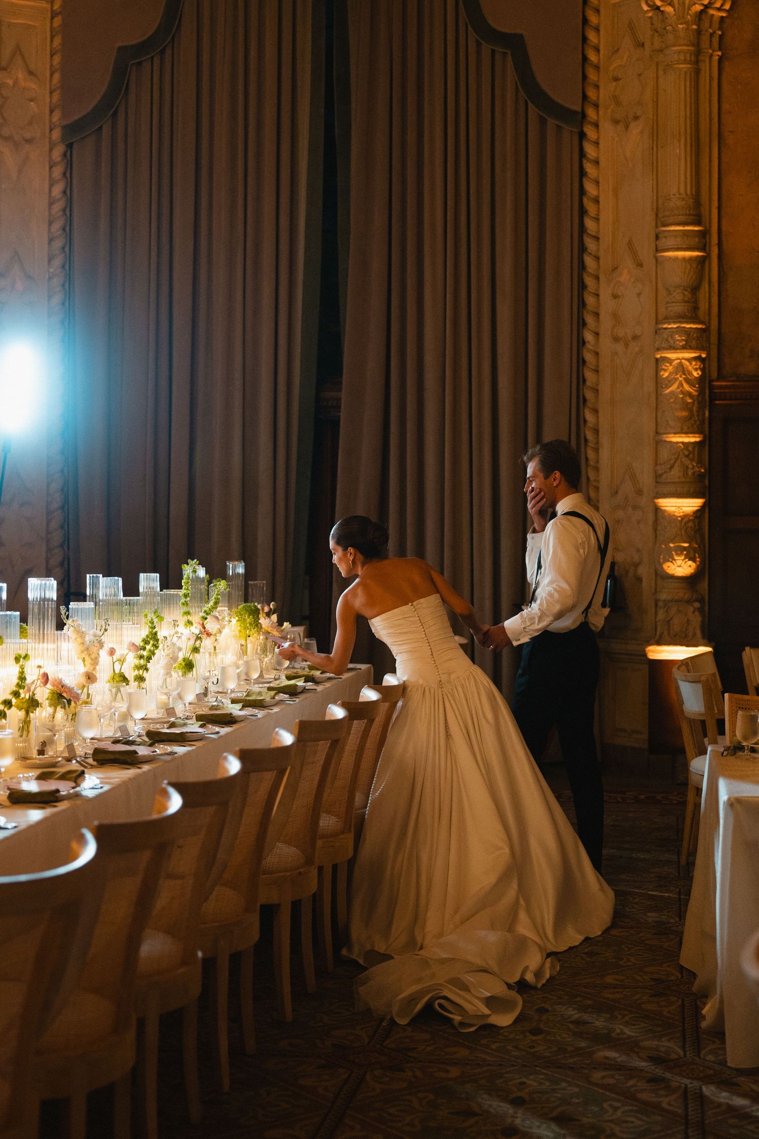 Bride and groom inspect elegantly set wedding reception table with candles and floral arrangements in ornate ballroom.