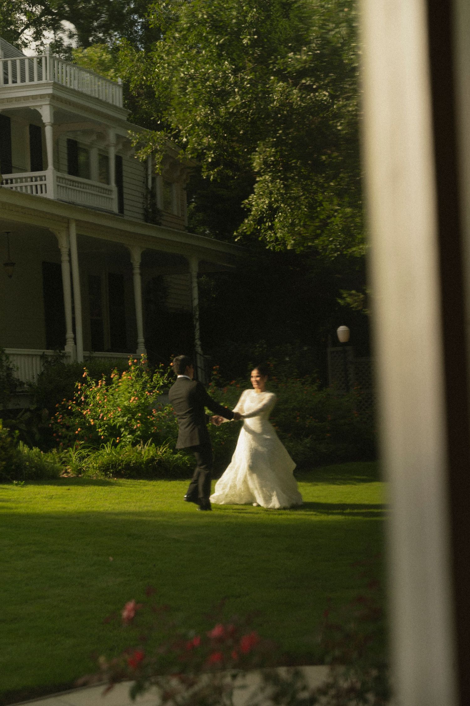 A bride in a flowing white wedding dress walks across a manicured lawn in front of a stately white mansion.