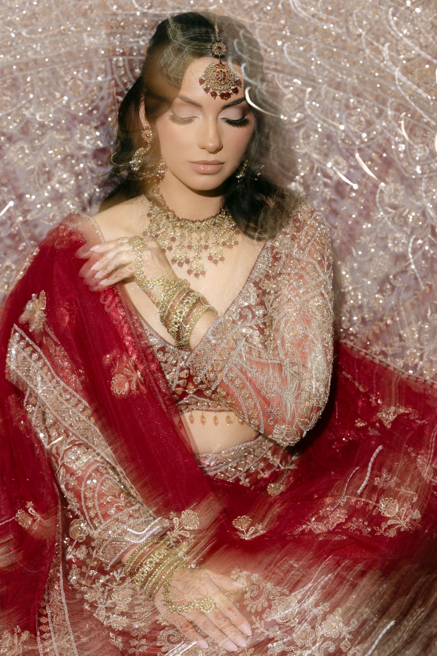 A traditional South Asian bride in a deep red lehenga adorned with intricate embroidery and gold jewelry poses elegantly.
