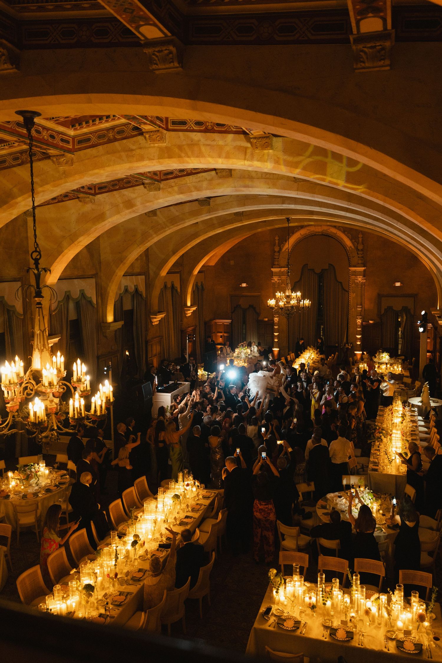 Elegant candlelit wedding reception in a grand ballroom with arched ceilings, long dining tables, and guests gathered on the dance floor.