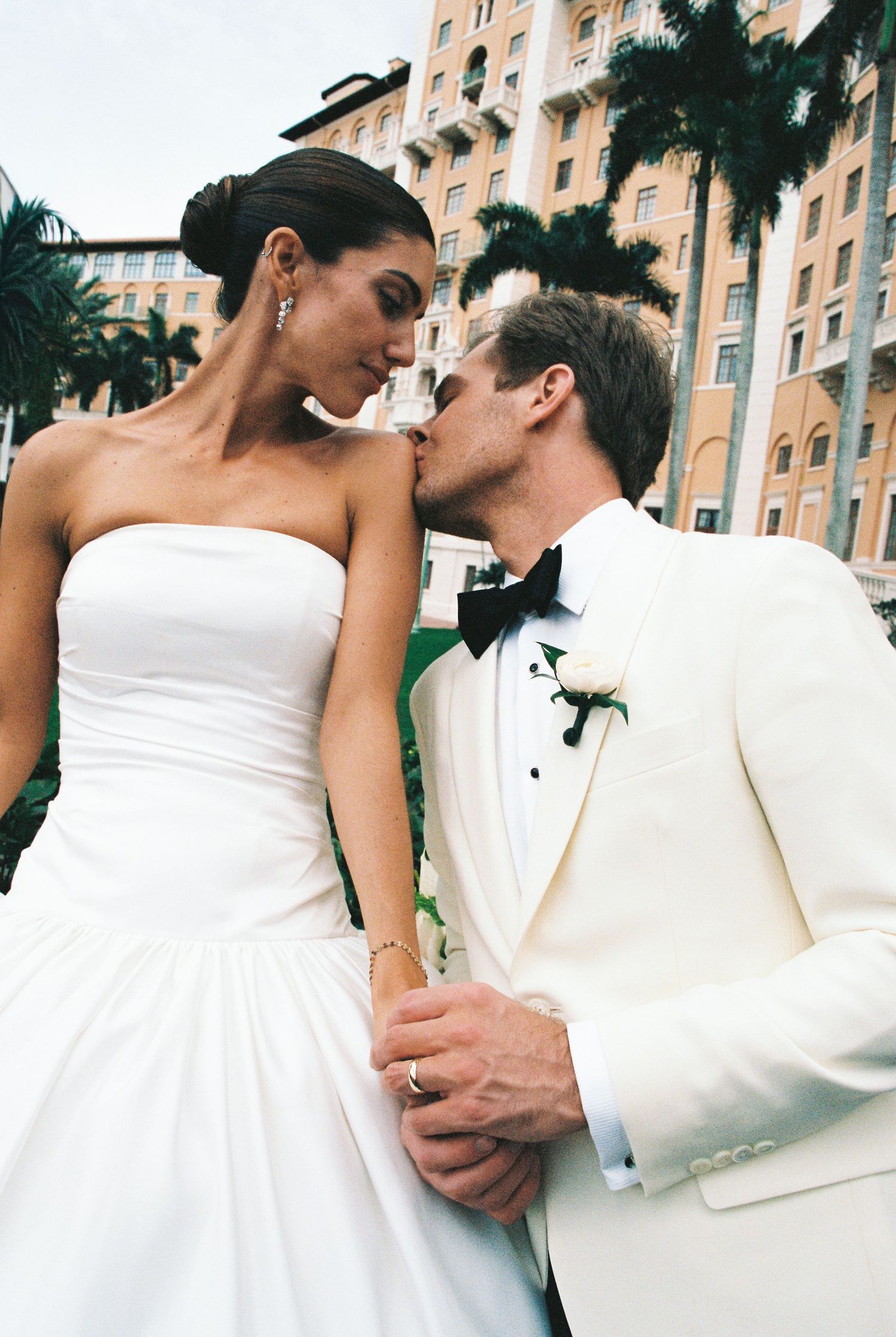 Groom in white tuxedo kisses bride on shoulder outside grand hotel surrounded by tall palm trees.