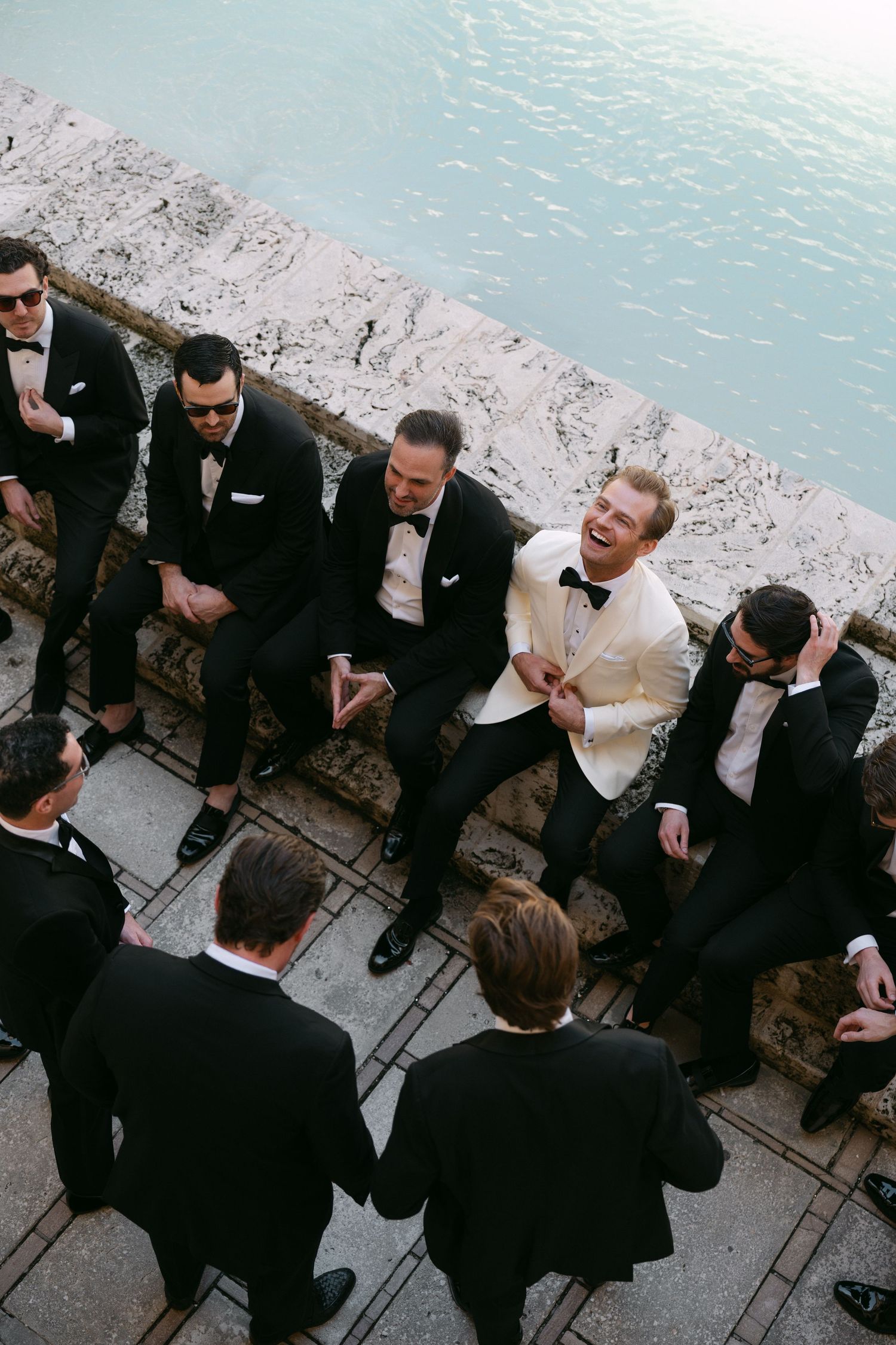Groom in white tuxedo jacket laughing with groomsmen in black tuxedos seated on stone steps near water.