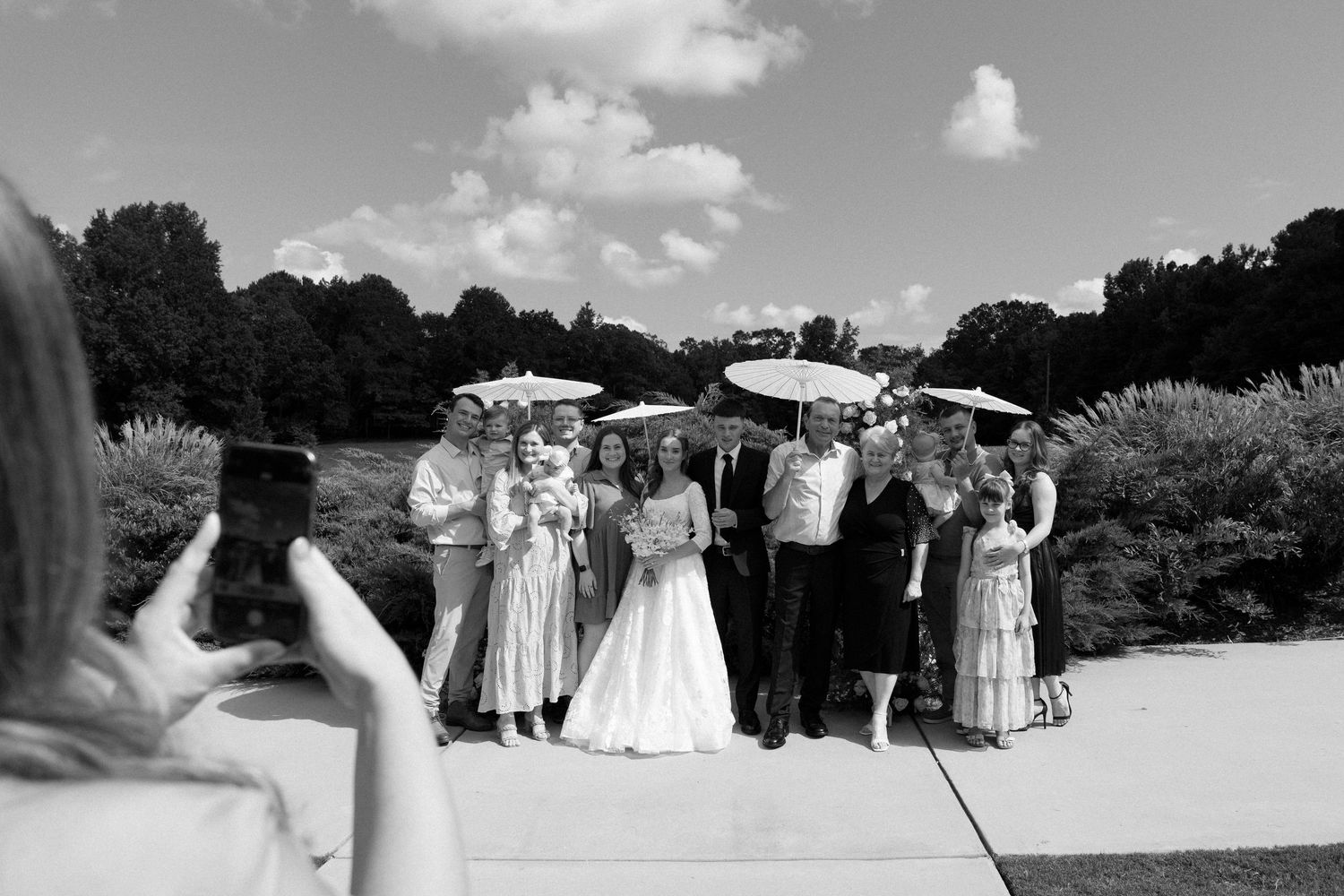 A black and white photo of a wedding party holding umbrellas while posing outdoors on a sunny day.