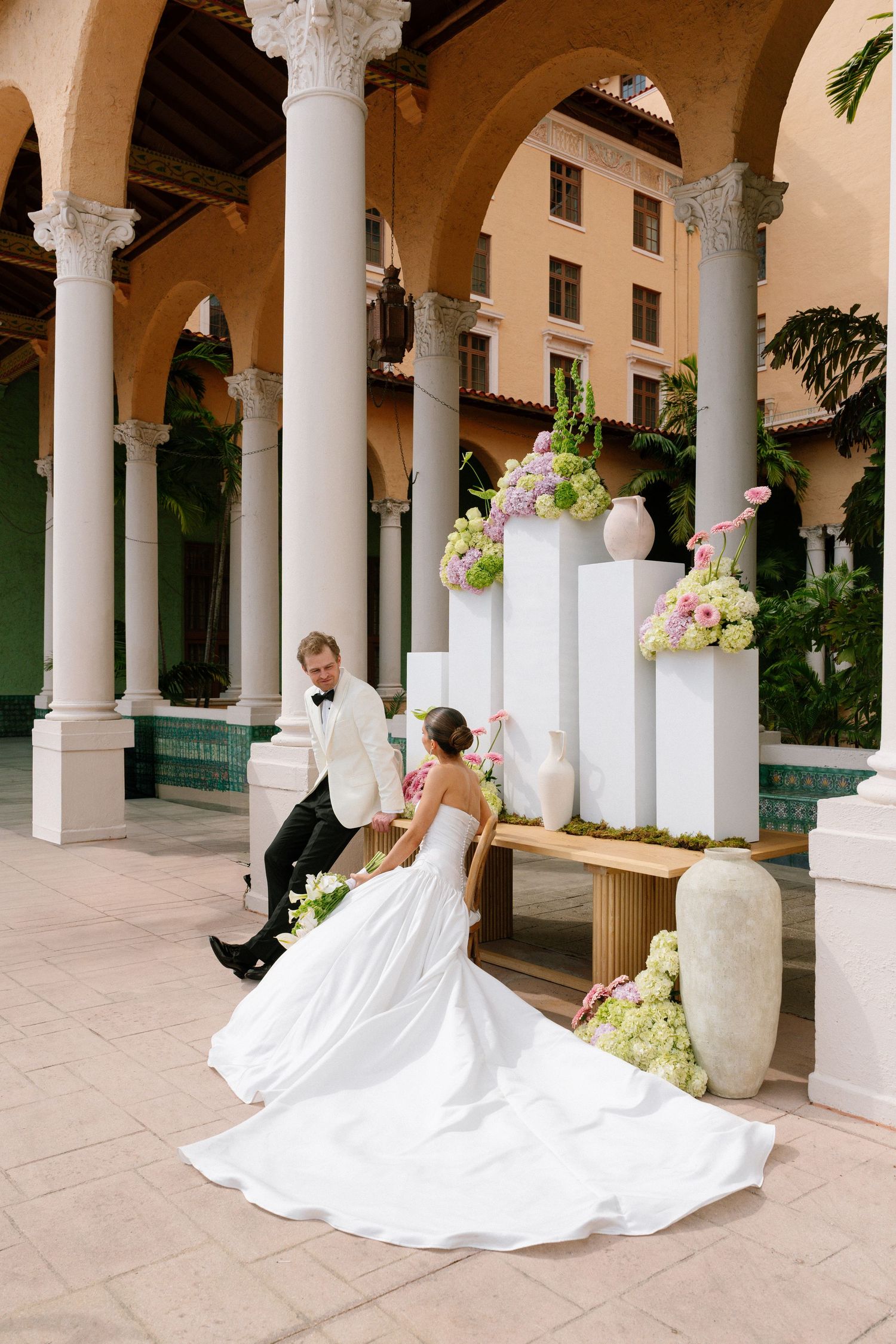 Bride in white gown and groom in tuxedo sit together in elegant columned courtyard with floral arrangements.