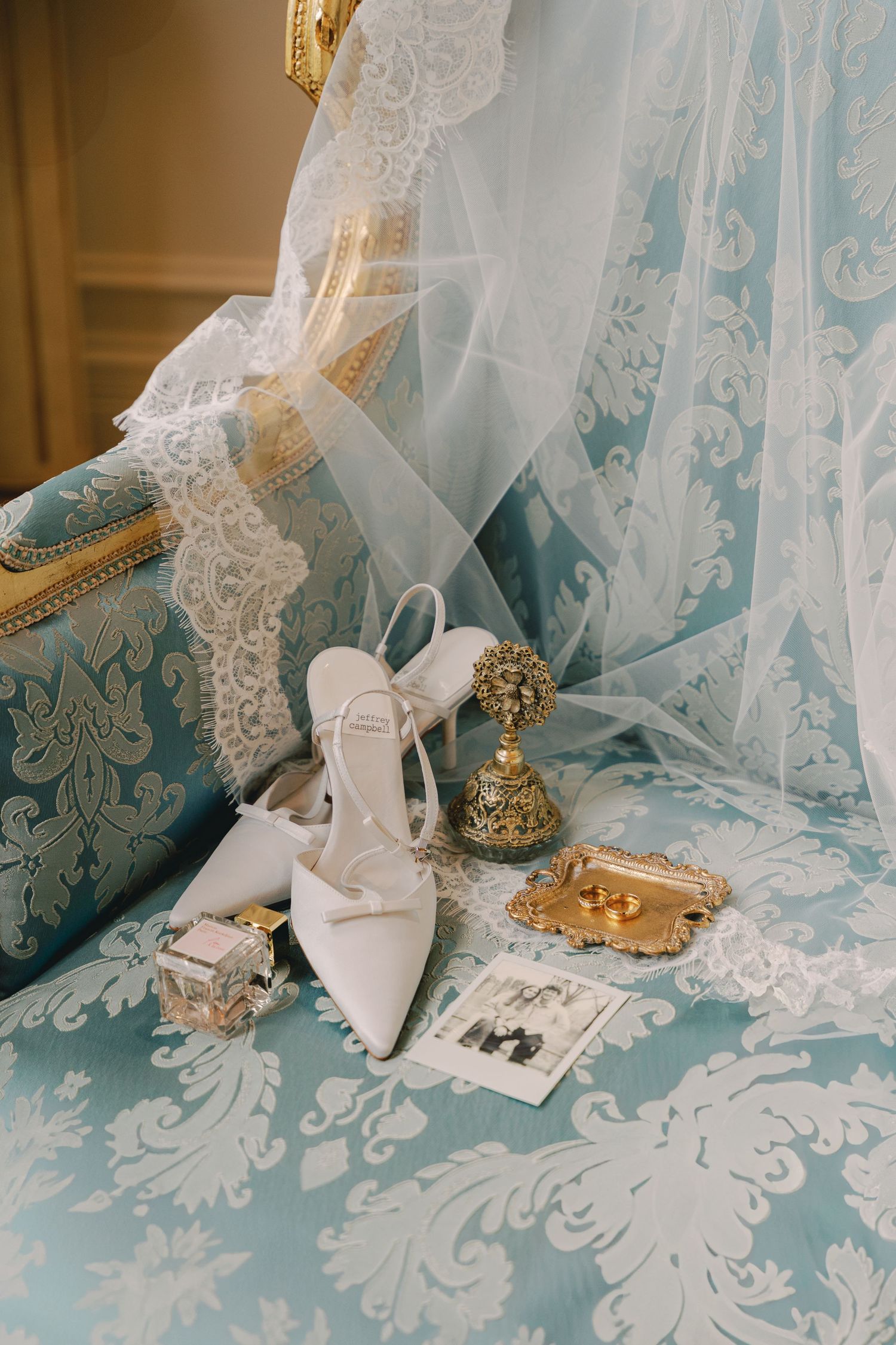 Wedding accessories including white heels and veil arranged on blue damask furniture.