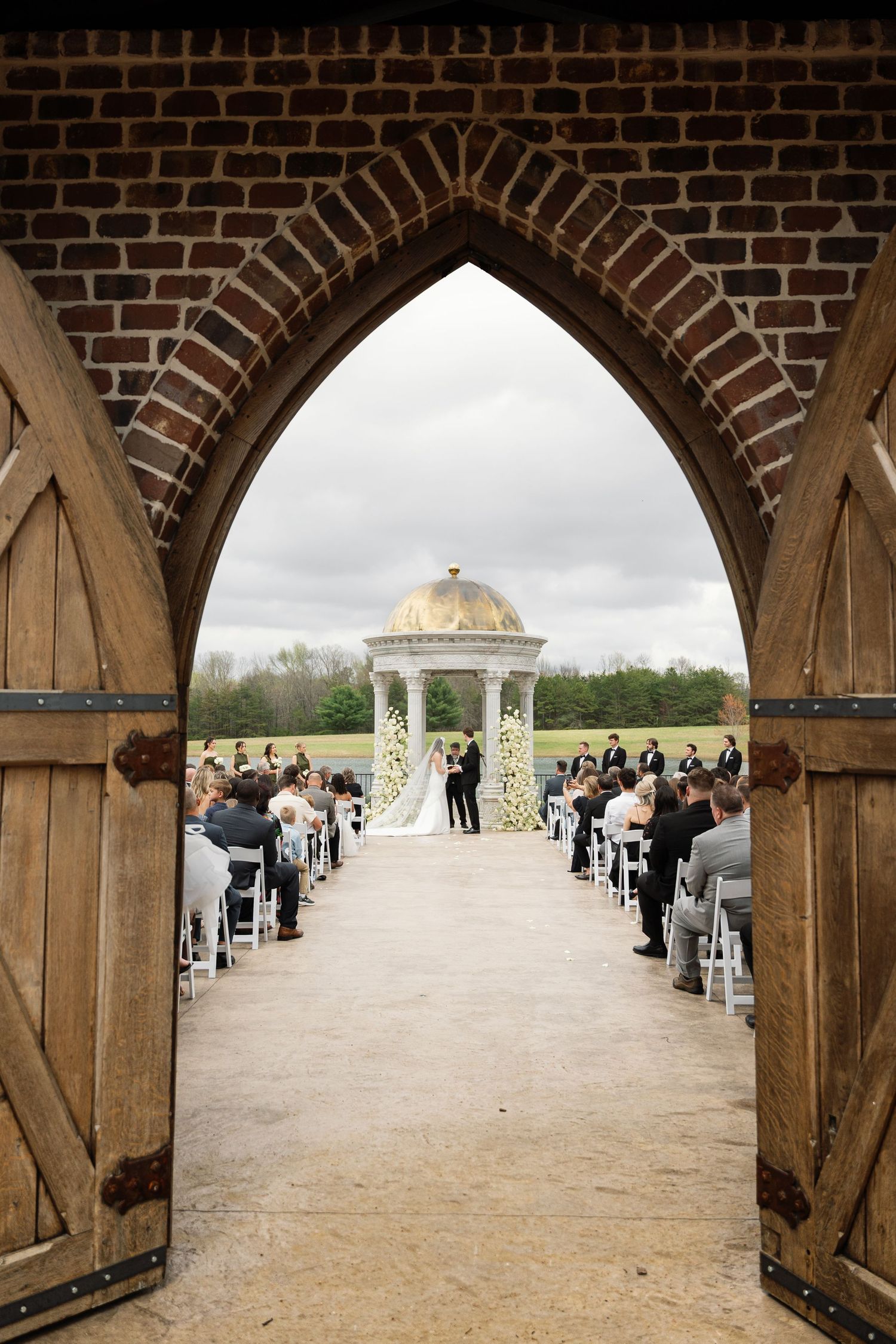 Outdoor wedding ceremony viewed through brick archway with guests seated before a domed gazebo on cloudy day.