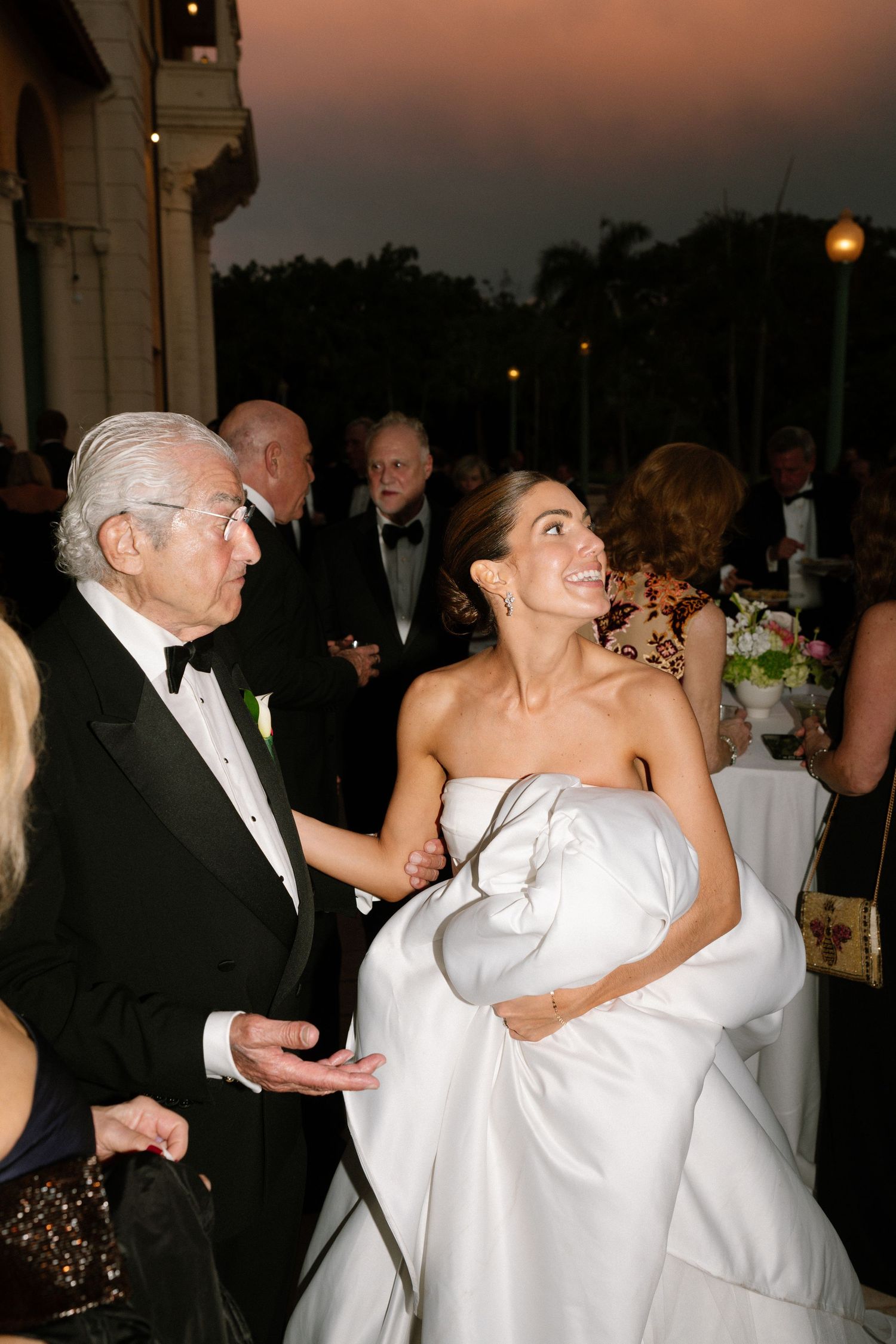 Bride in strapless white gown with wrap smiles at elderly guest in tuxedo during outdoor evening wedding reception.