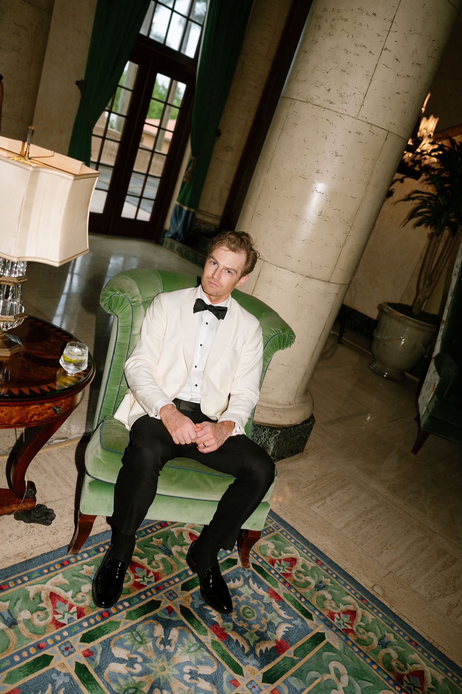 Groom in white tuxedo jacket and bow tie sitting in green velvet chair in elegant marble lobby.