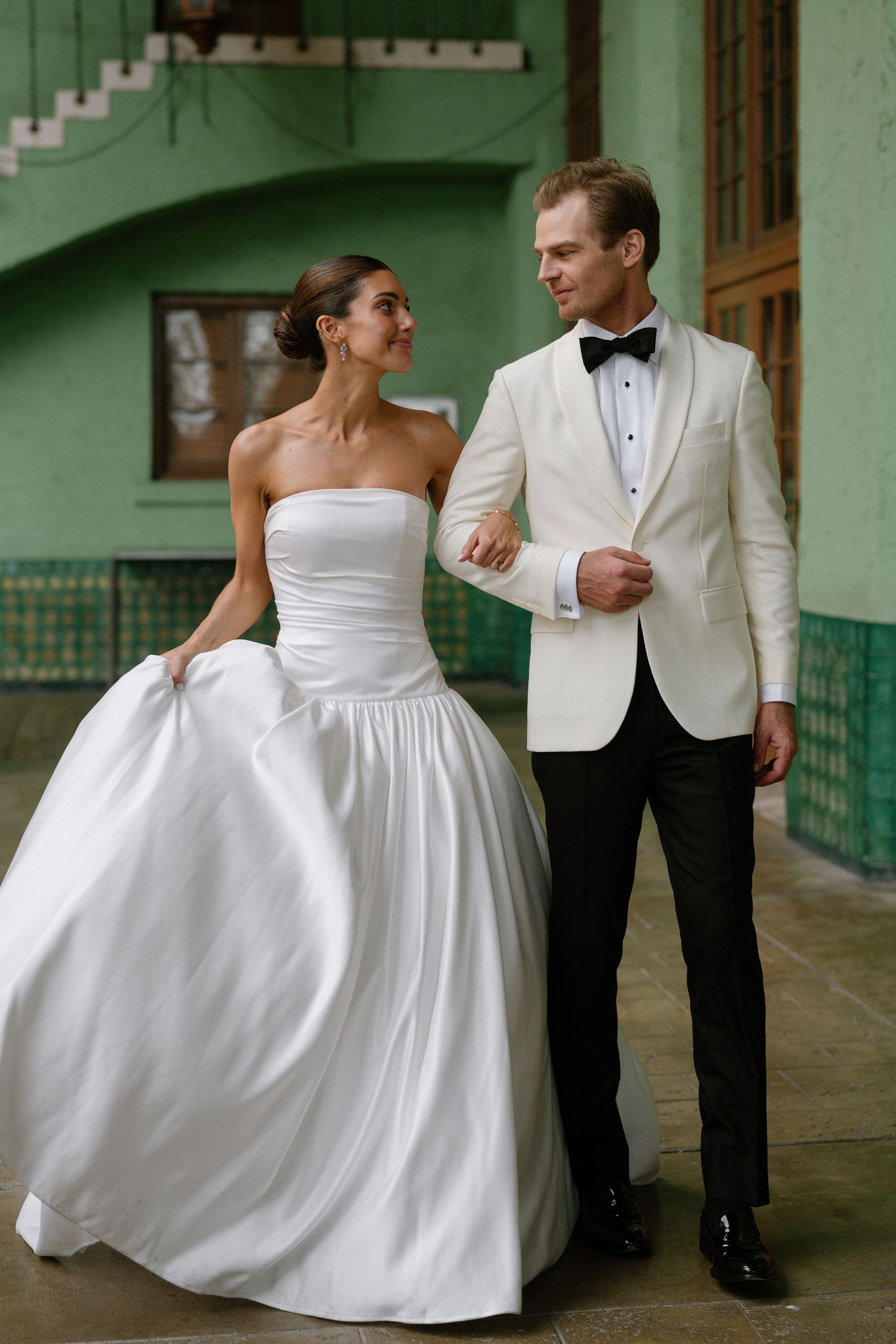 Bride and groom posing together, she in strapless white ballgown, he in white tuxedo jacket with black bow tie.