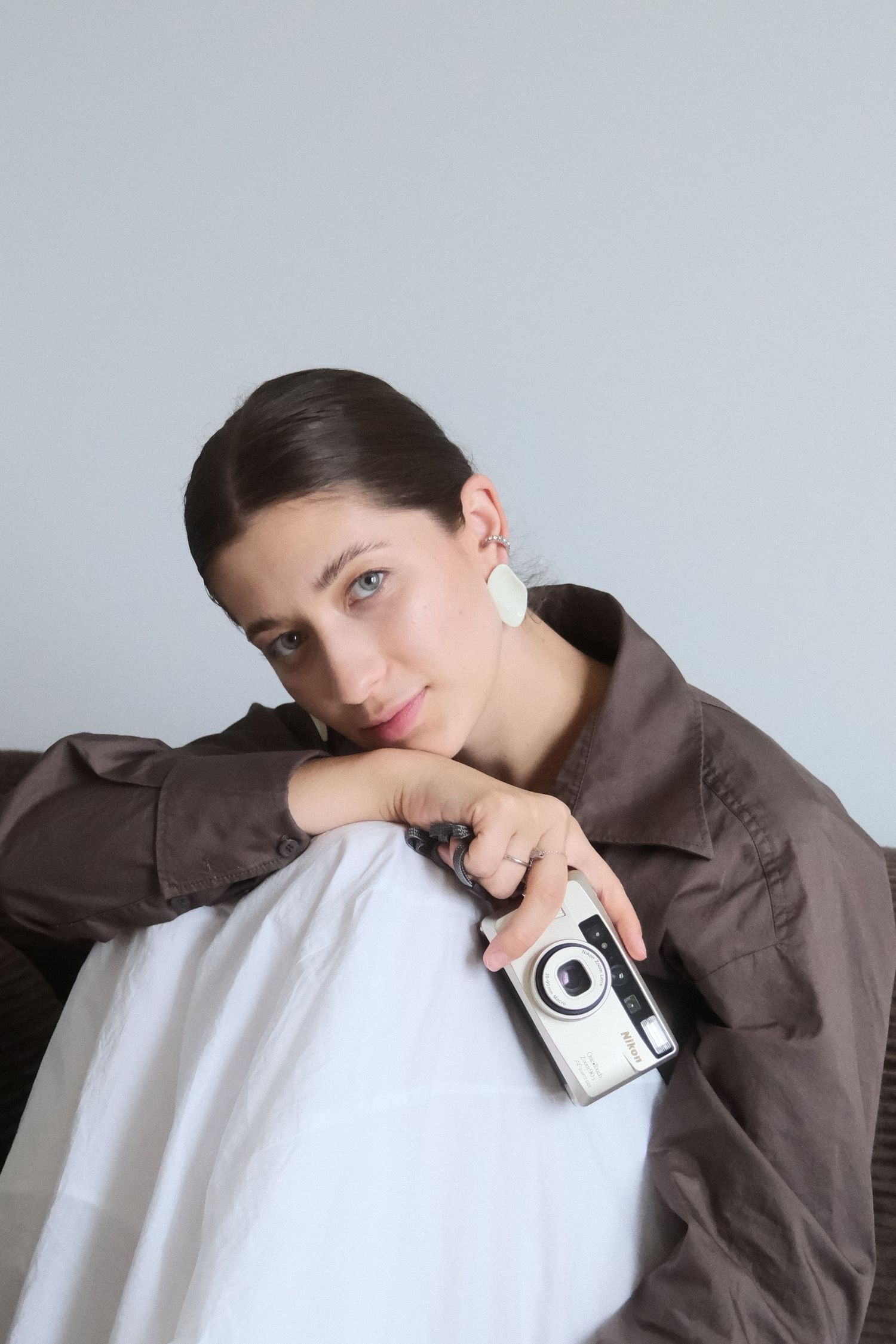 A young woman in a brown shirt leans on a white surface, holding a vintage camera, wearing statement earrings.
