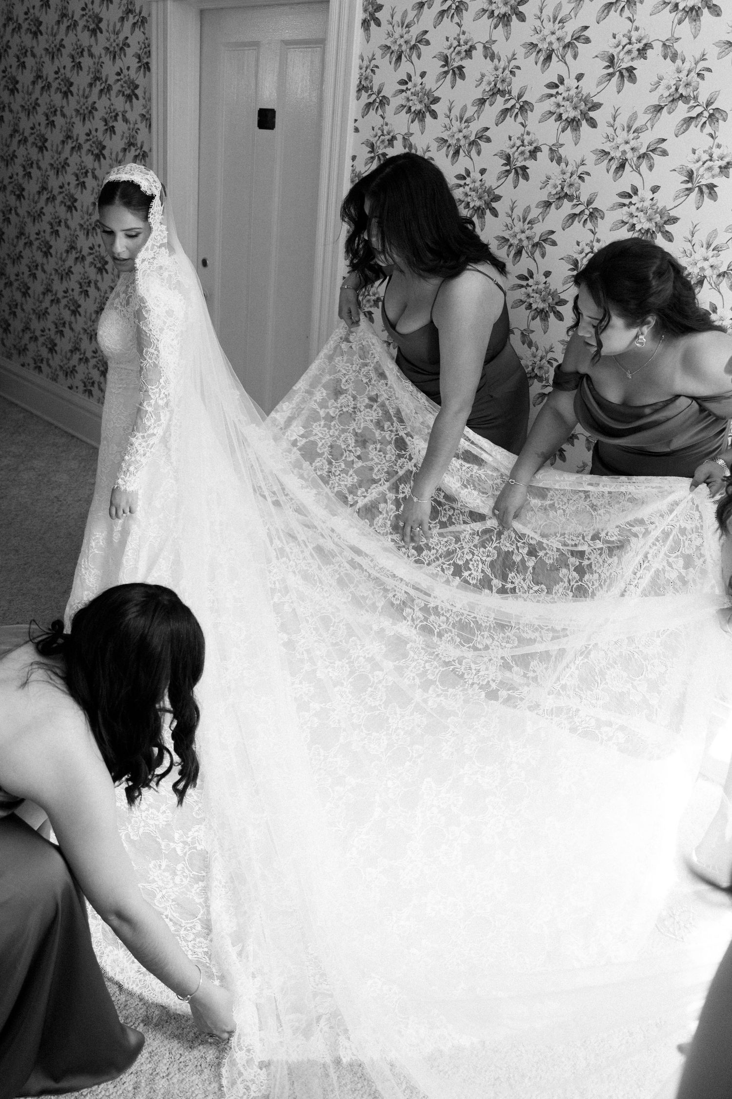 Bridesmaids help arrange a delicate lace wedding veil against floral wallpaper in black and white photograph.