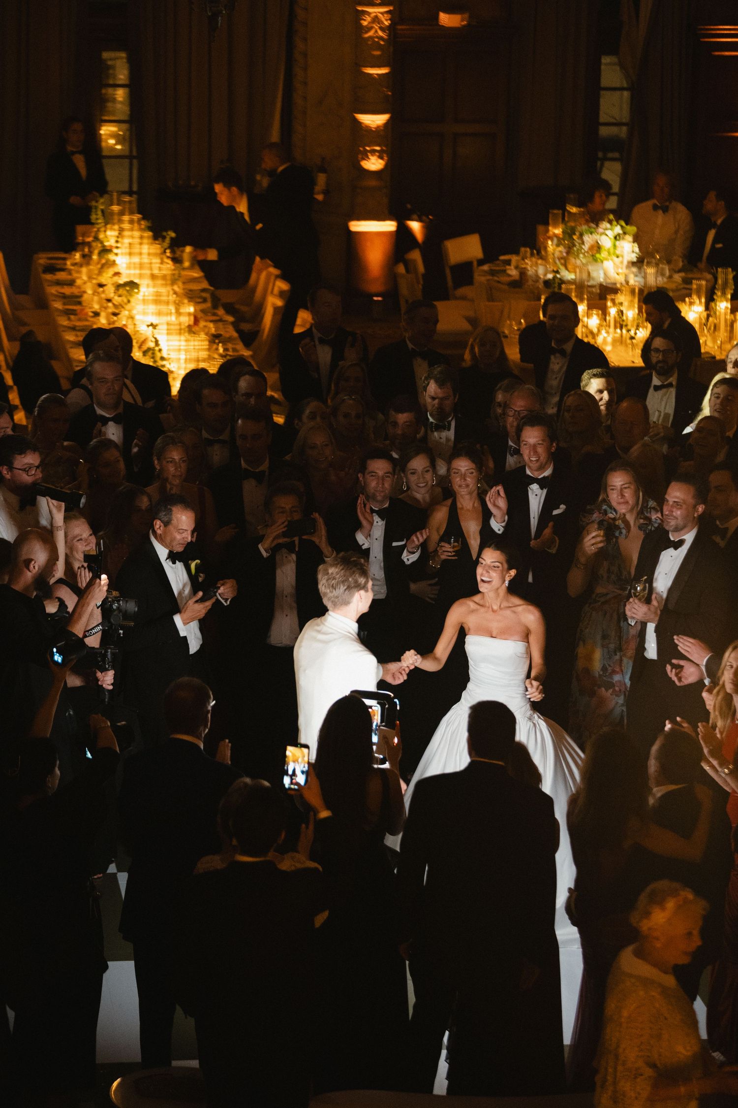 Bride and groom enter their wedding reception to cheering guests in a warmly lit elegant ballroom.