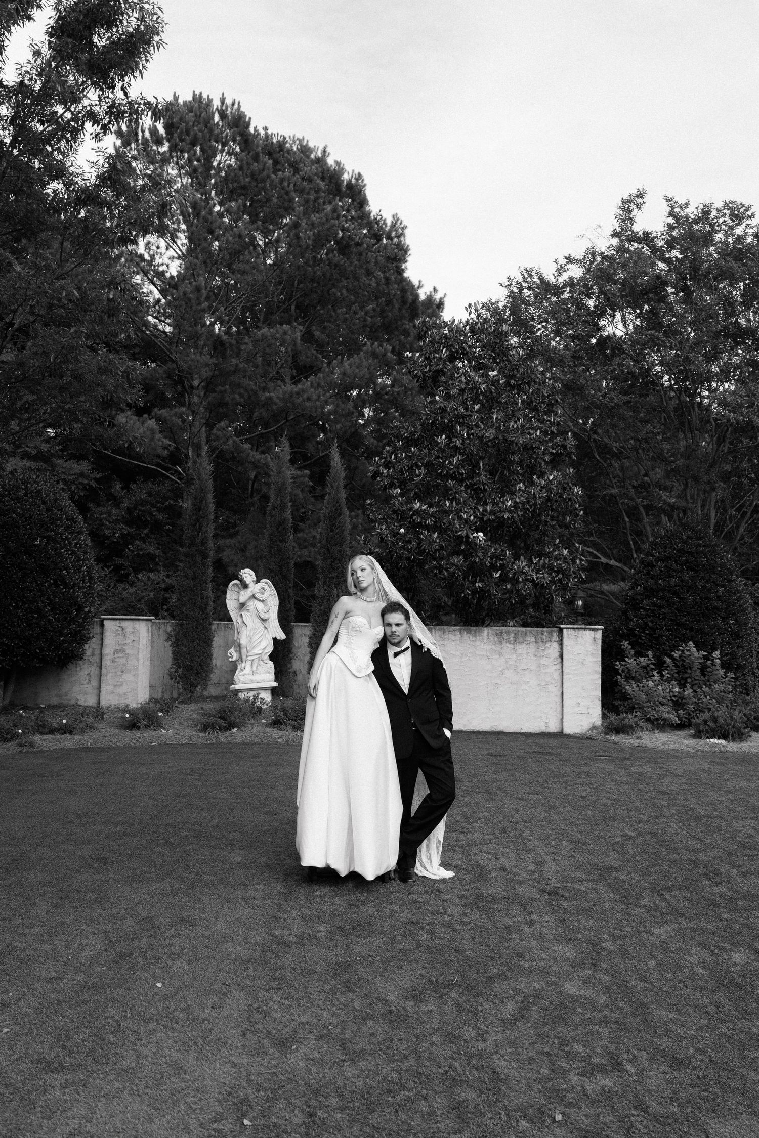 Black and white wedding photo of bride and groom posing in garden with angel statue and trees.