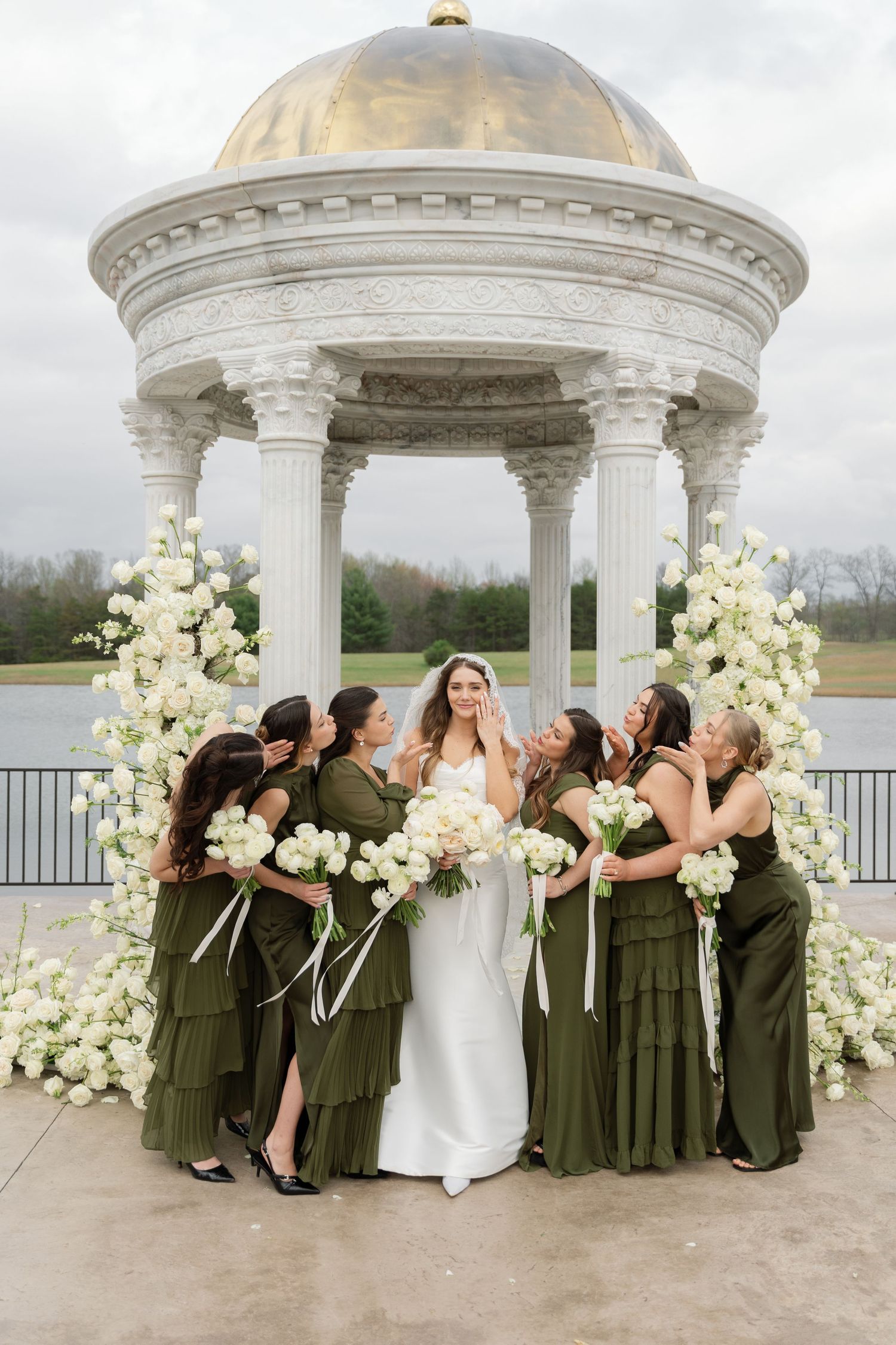 A bridal party in olive green dresses gathers under an elegant white gazebo decorated with cream florals.