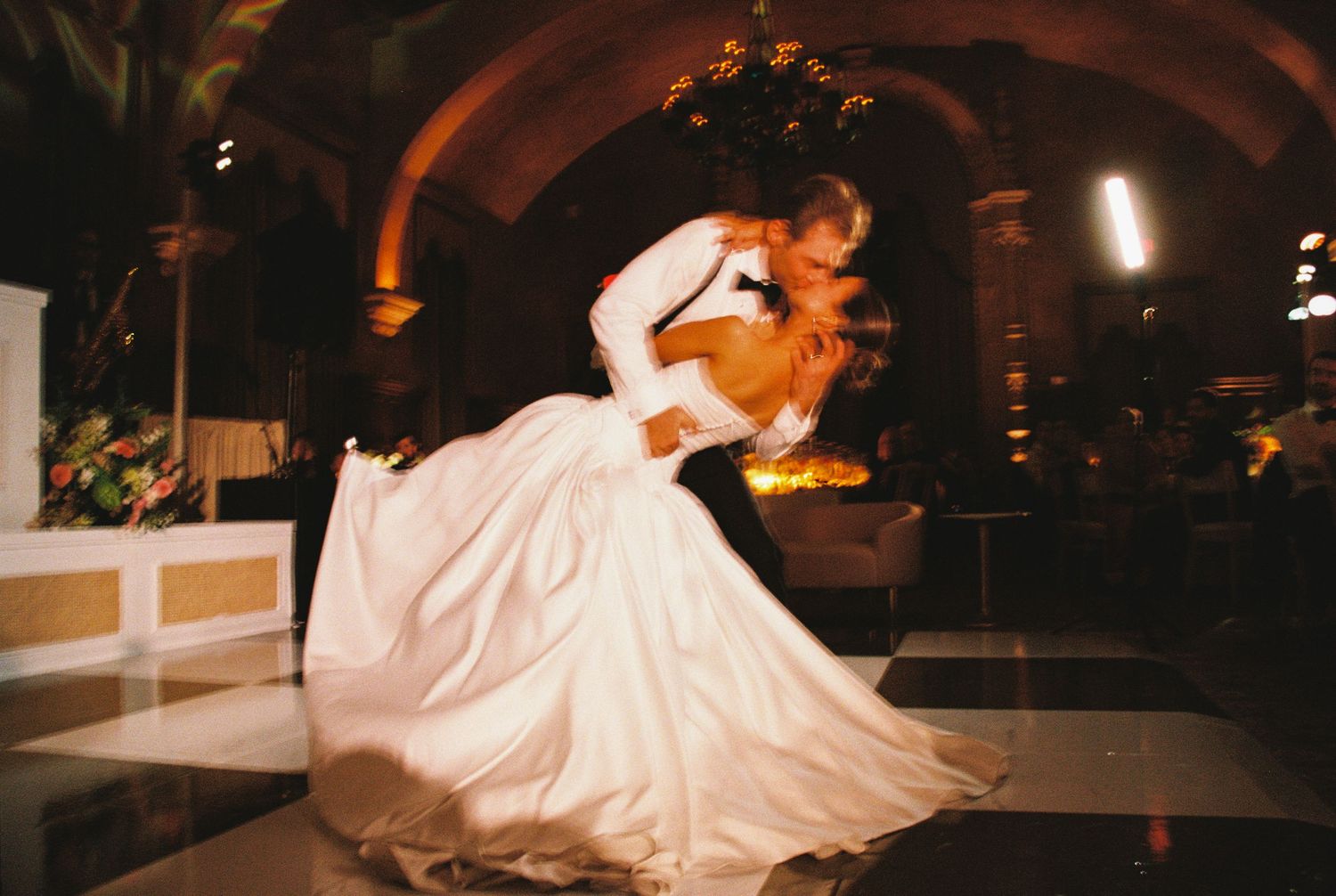Bride and groom share a romantic dip dance on the wedding dance floor in an elegant venue with arched columns.