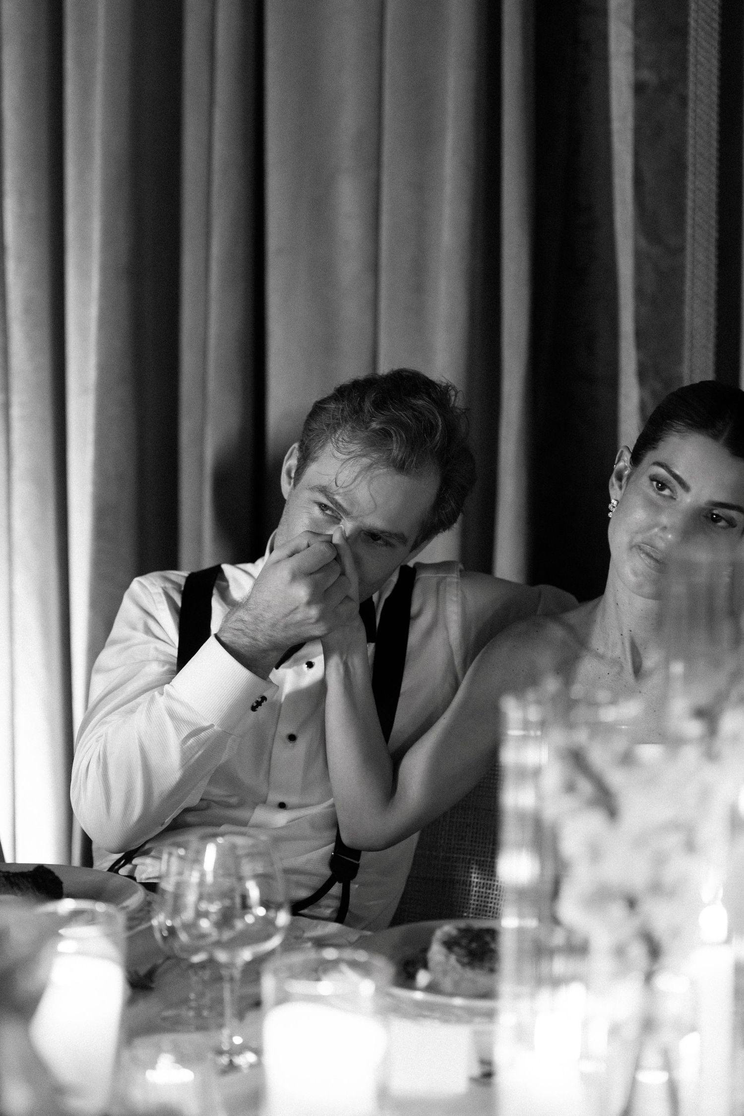 Black and white photo of emotional groom wiping tears at wedding reception table while bride comforts him.