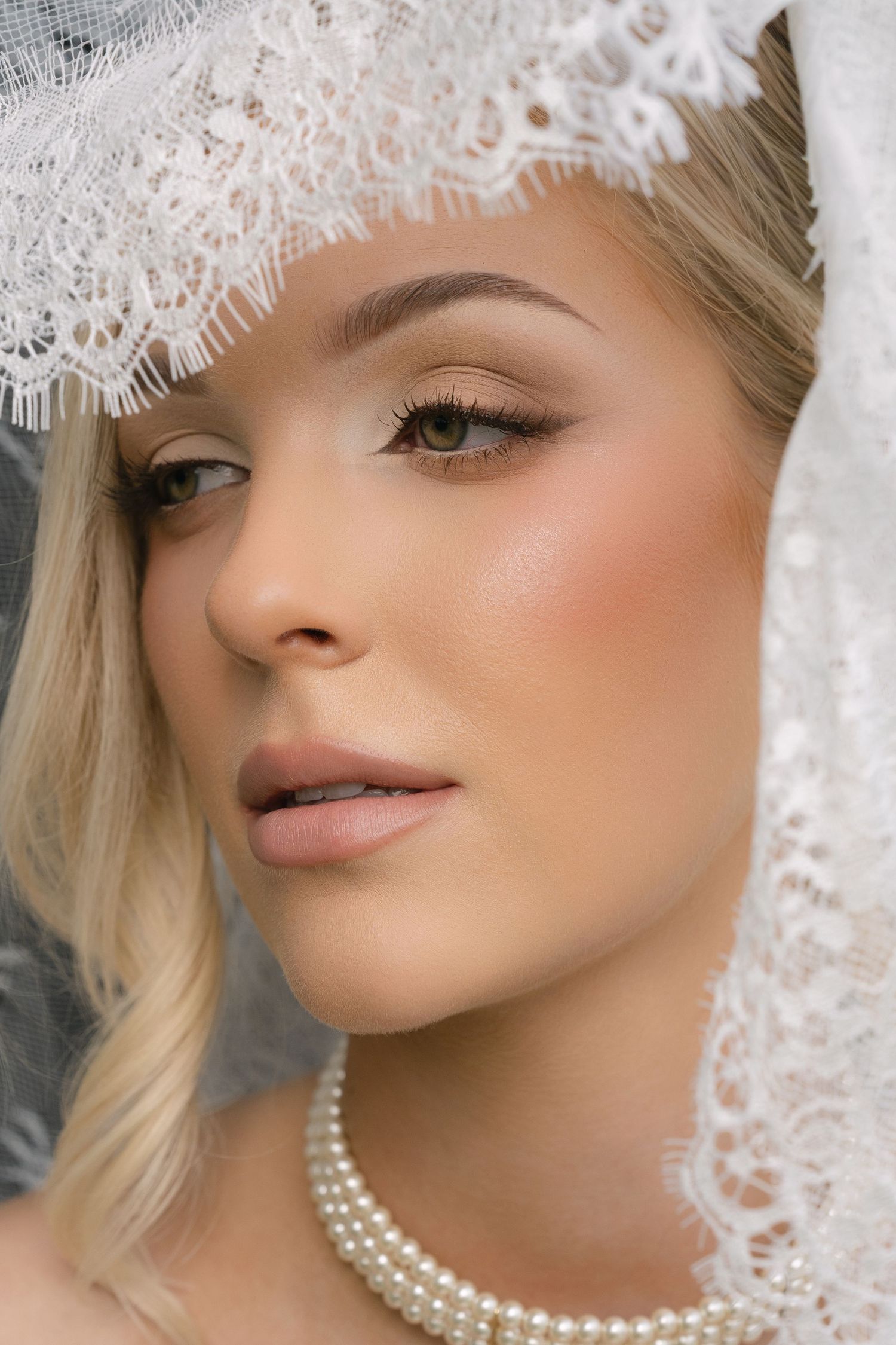 Blonde bride wearing lace veil and pearl necklace with elegant bridal makeup including winged eyeliner and rosy blush.
