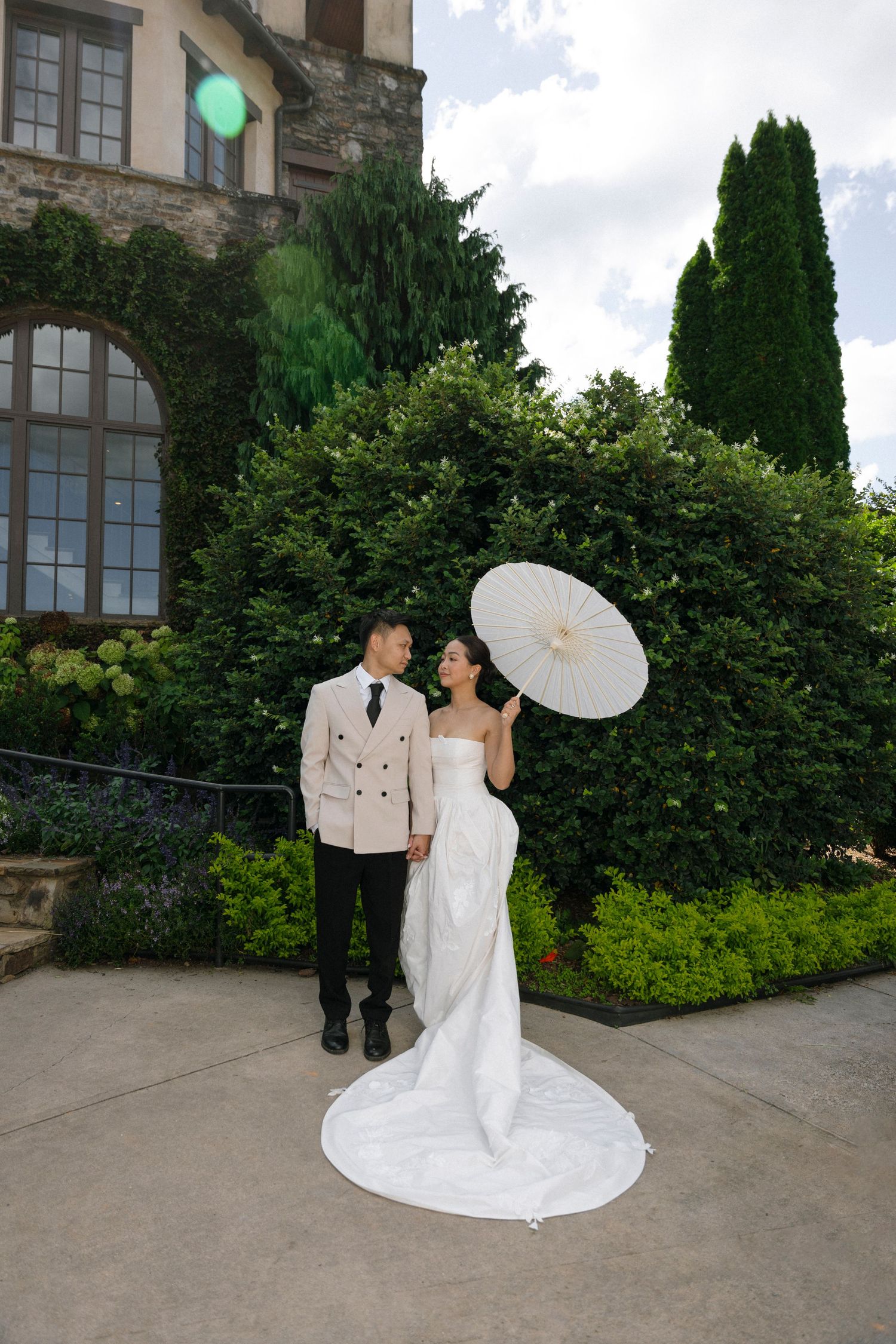A couple poses for a wedding photo against an ivy-covered building with the bride holding a white parasol.