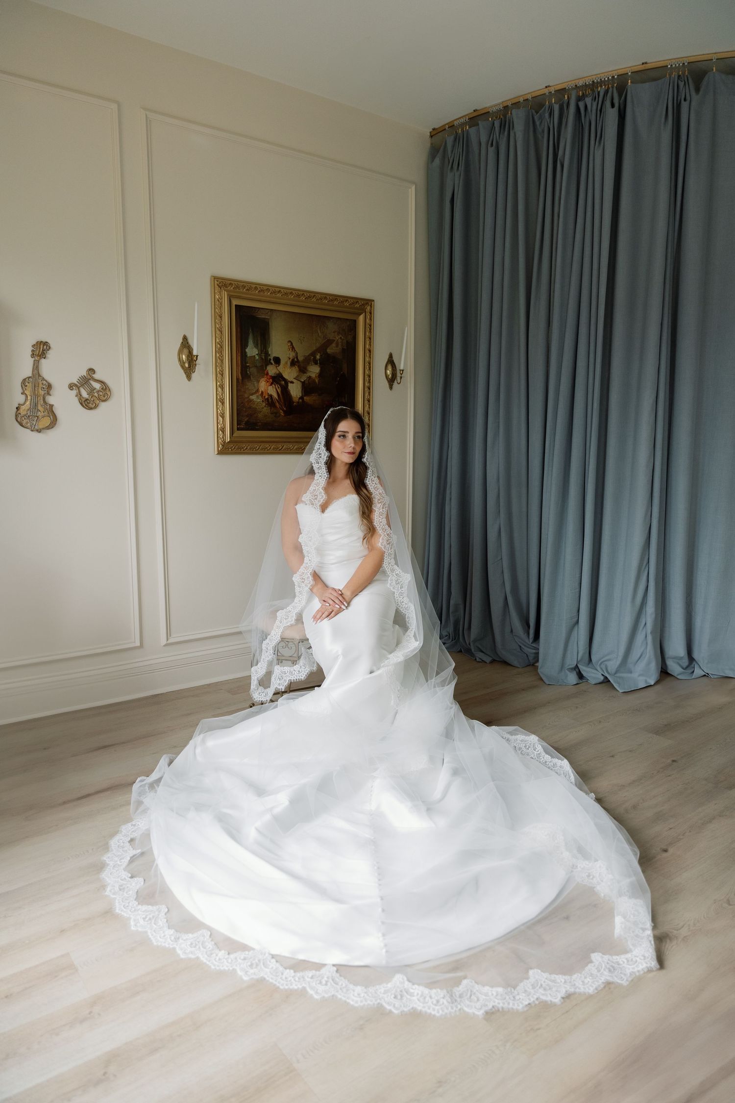 A bride in an elegant white lace wedding gown with a long train poses in a room with blue curtains and wall decor.