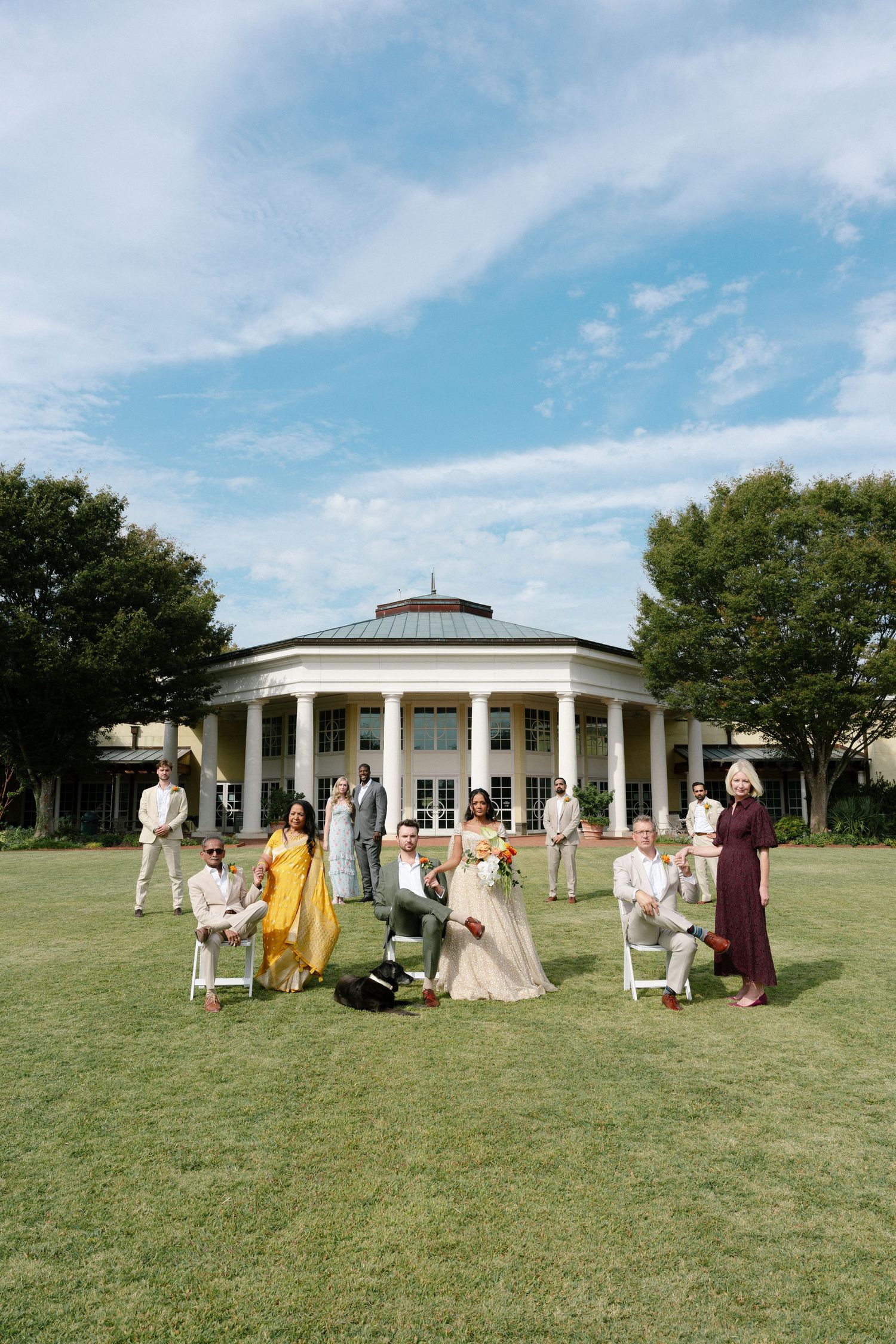 Wedding party gathers on a sunny day outside a white columned pavilion on a green lawn.