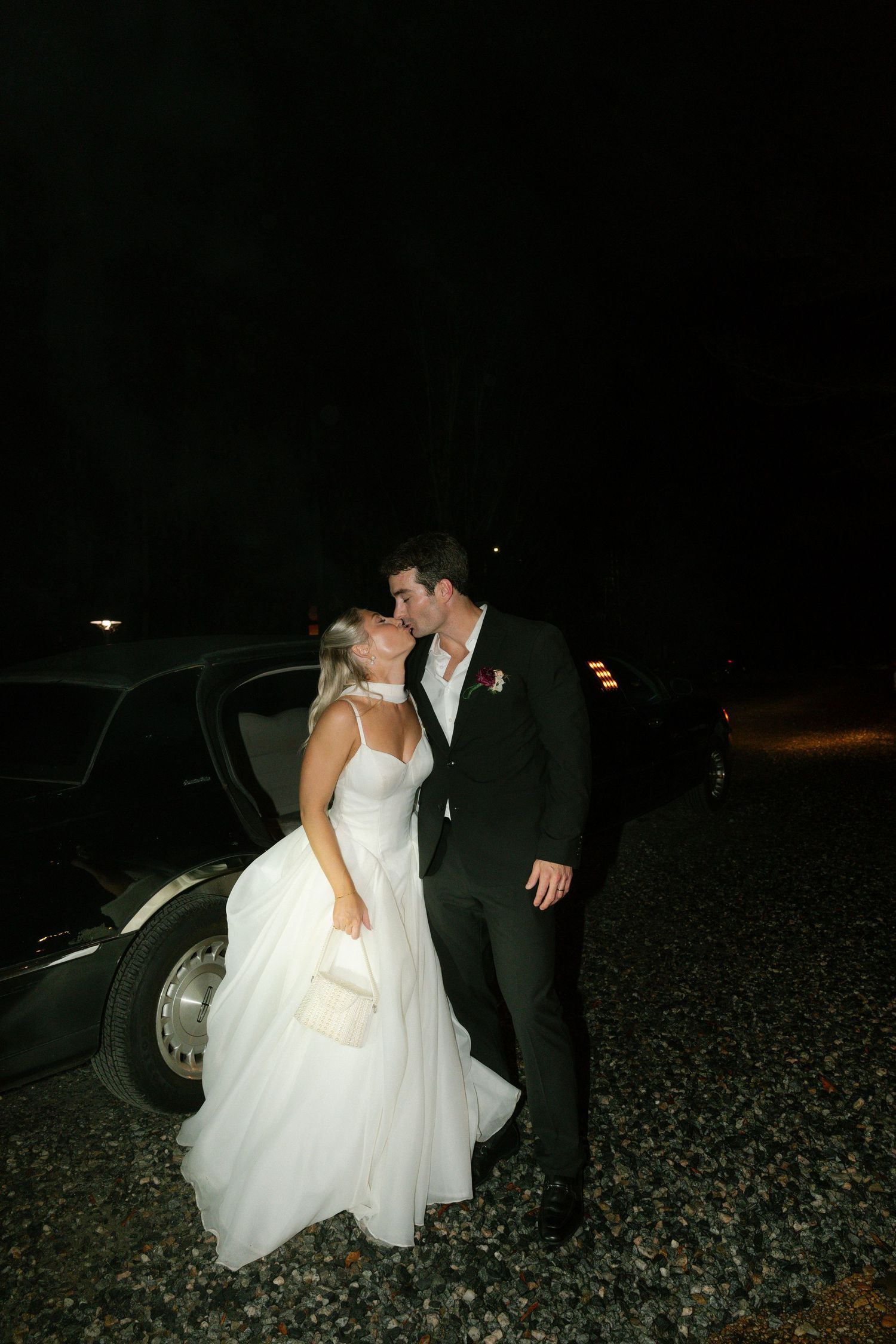 A couple in wedding attire share a romantic moment next to a car at night.