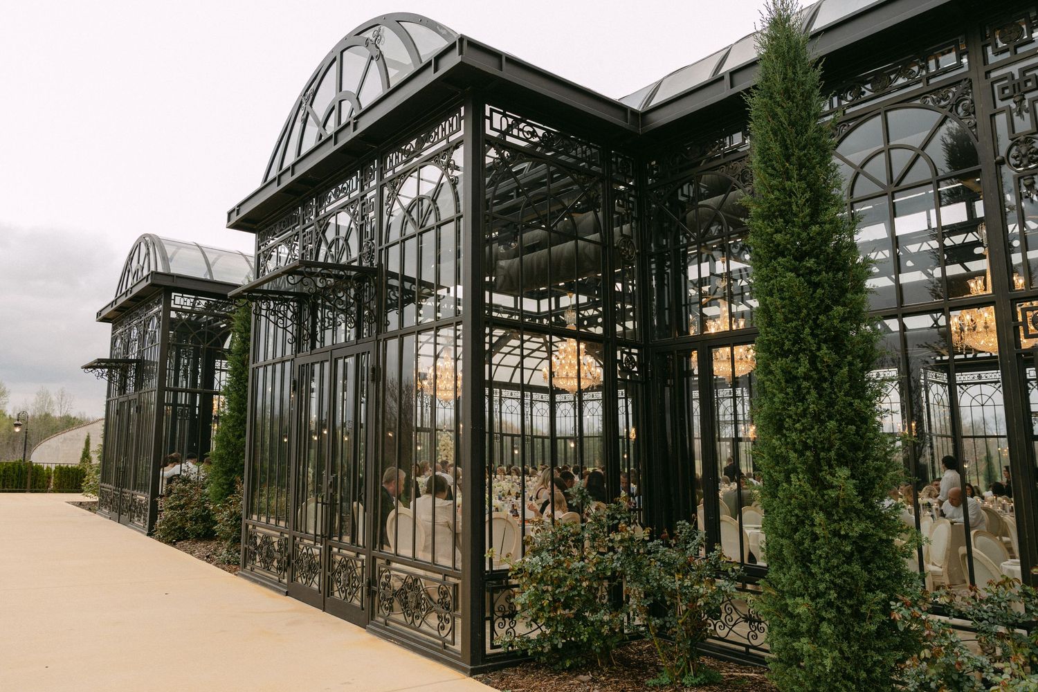 An elegant Victorian-style greenhouse with ornate black metal and glass architecture stands beside tall cypress trees.