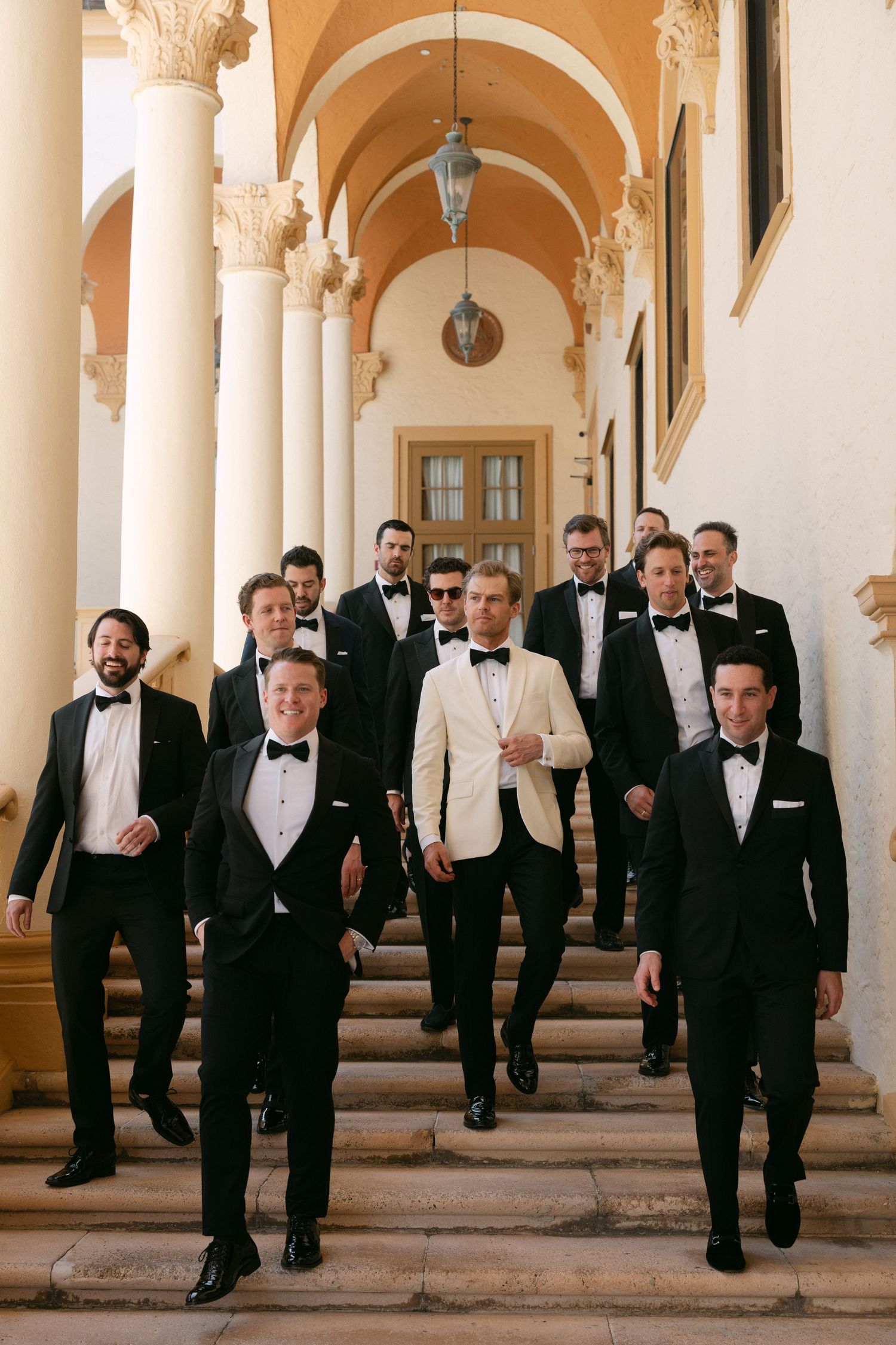 Groomsmen in black tuxedos descend grand stone steps at elegant venue, led by groom in white dinner jacket.