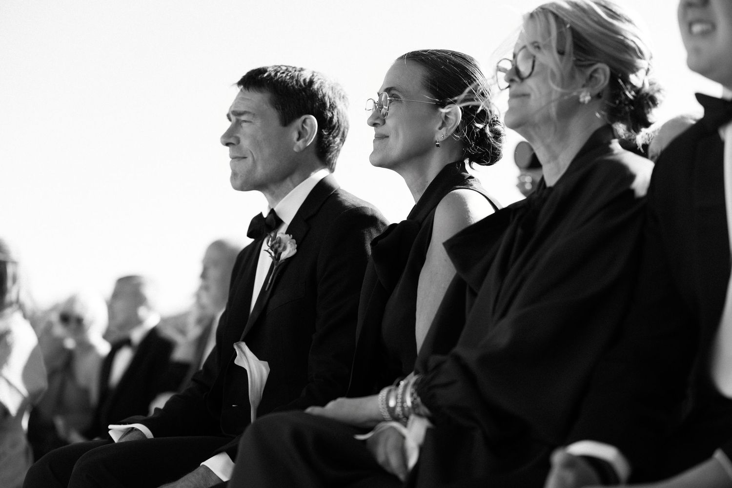 Black and white photo of elegantly dressed wedding guests seated and smiling during an outdoor ceremony.