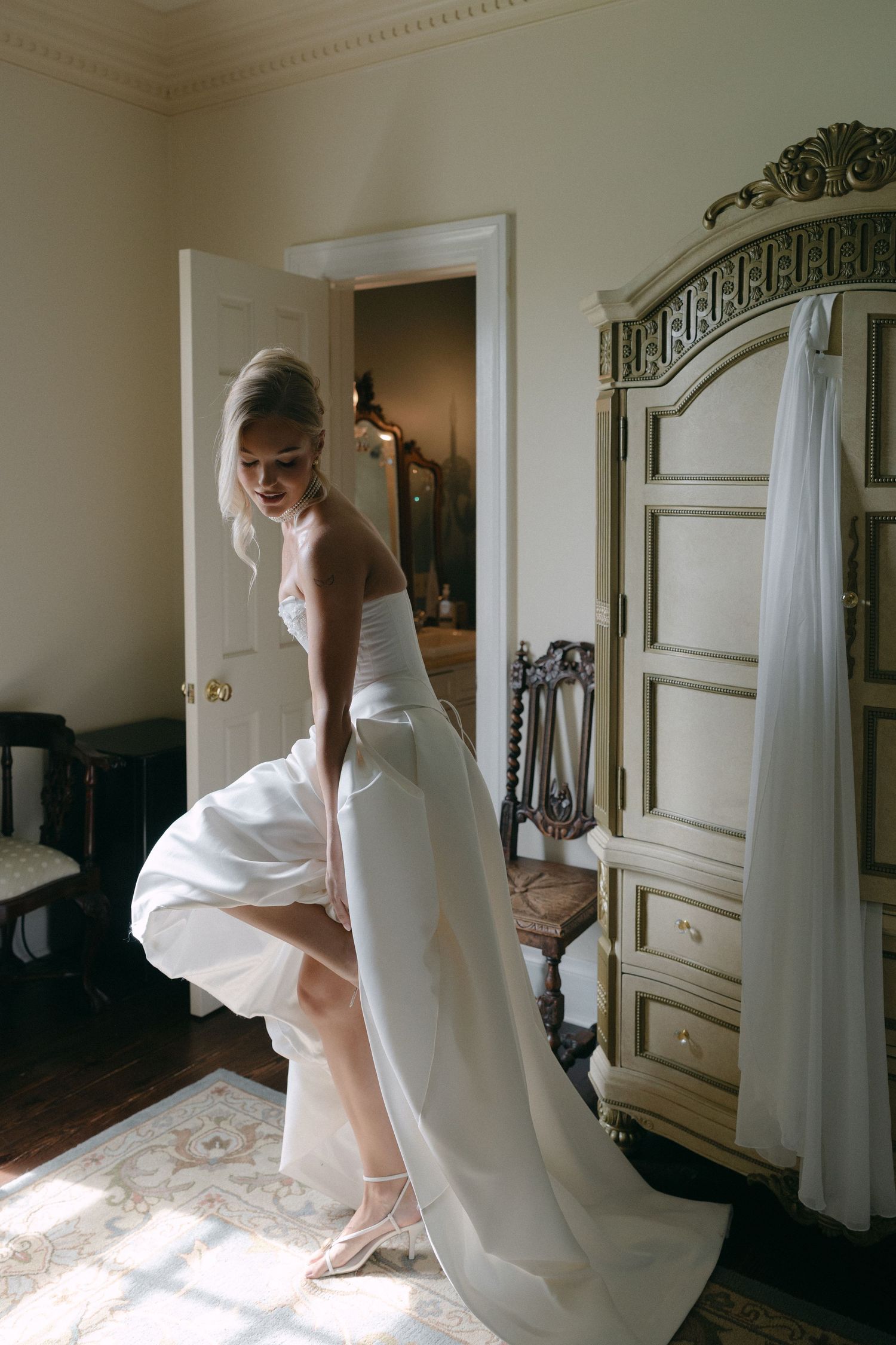 Bride in a flowing white strapless wedding gown lifting her skirt in an elegant vintage bedroom with ornate armoire.