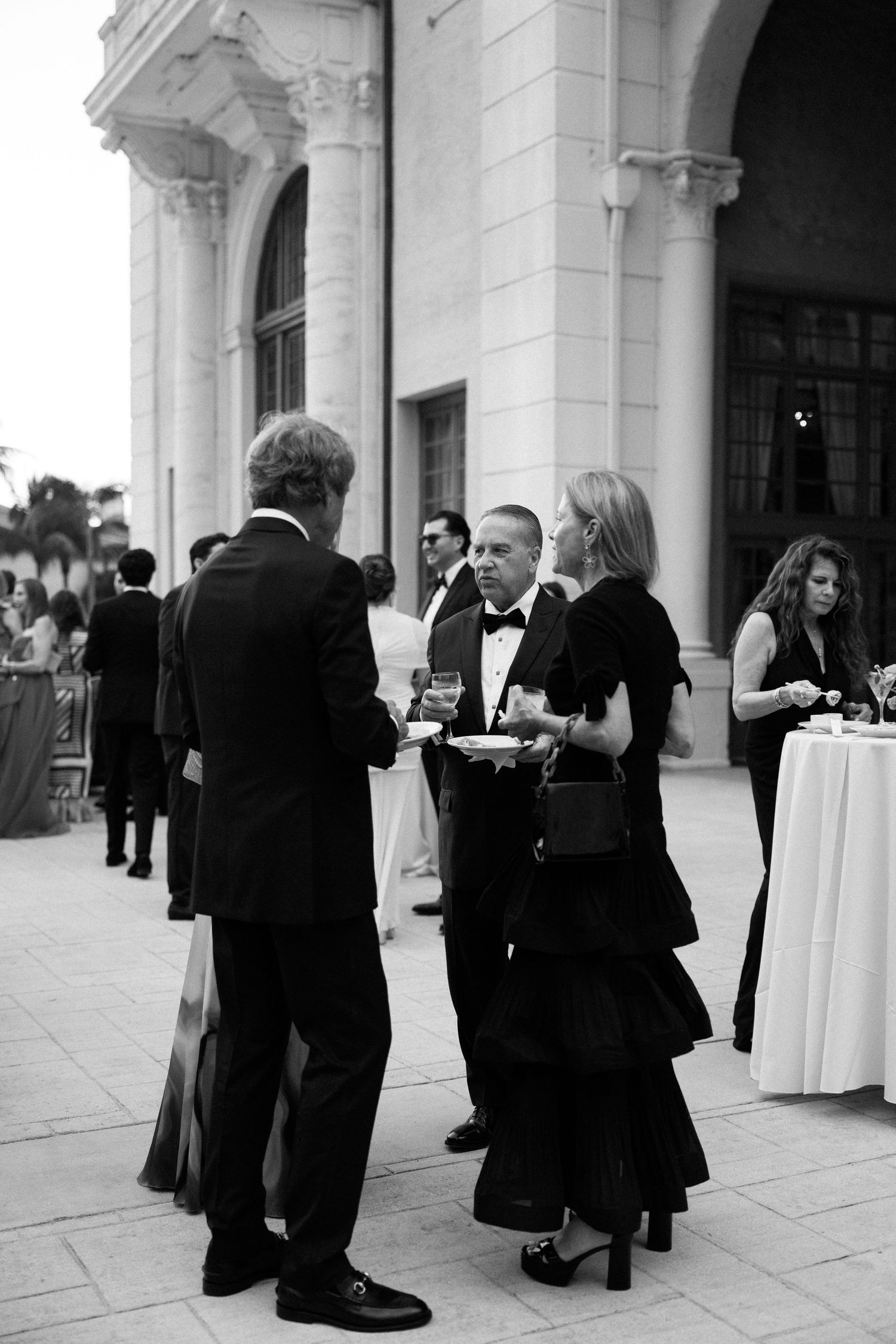 Elegantly dressed guests mingle outdoors at a formal black-tie event near a grand classical building.