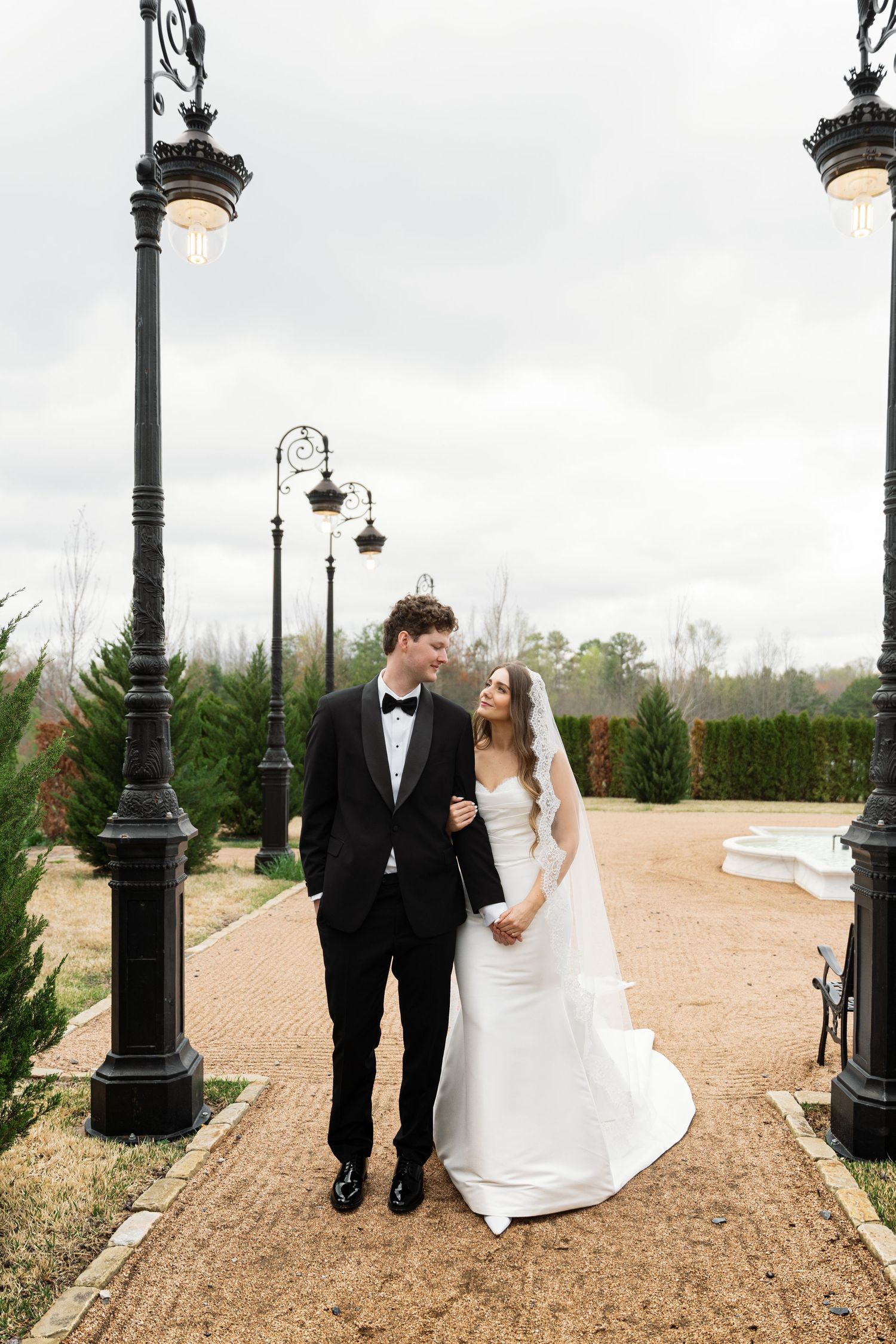 A romantic wedding couple poses on a garden path between decorative lamp posts on an overcast day.