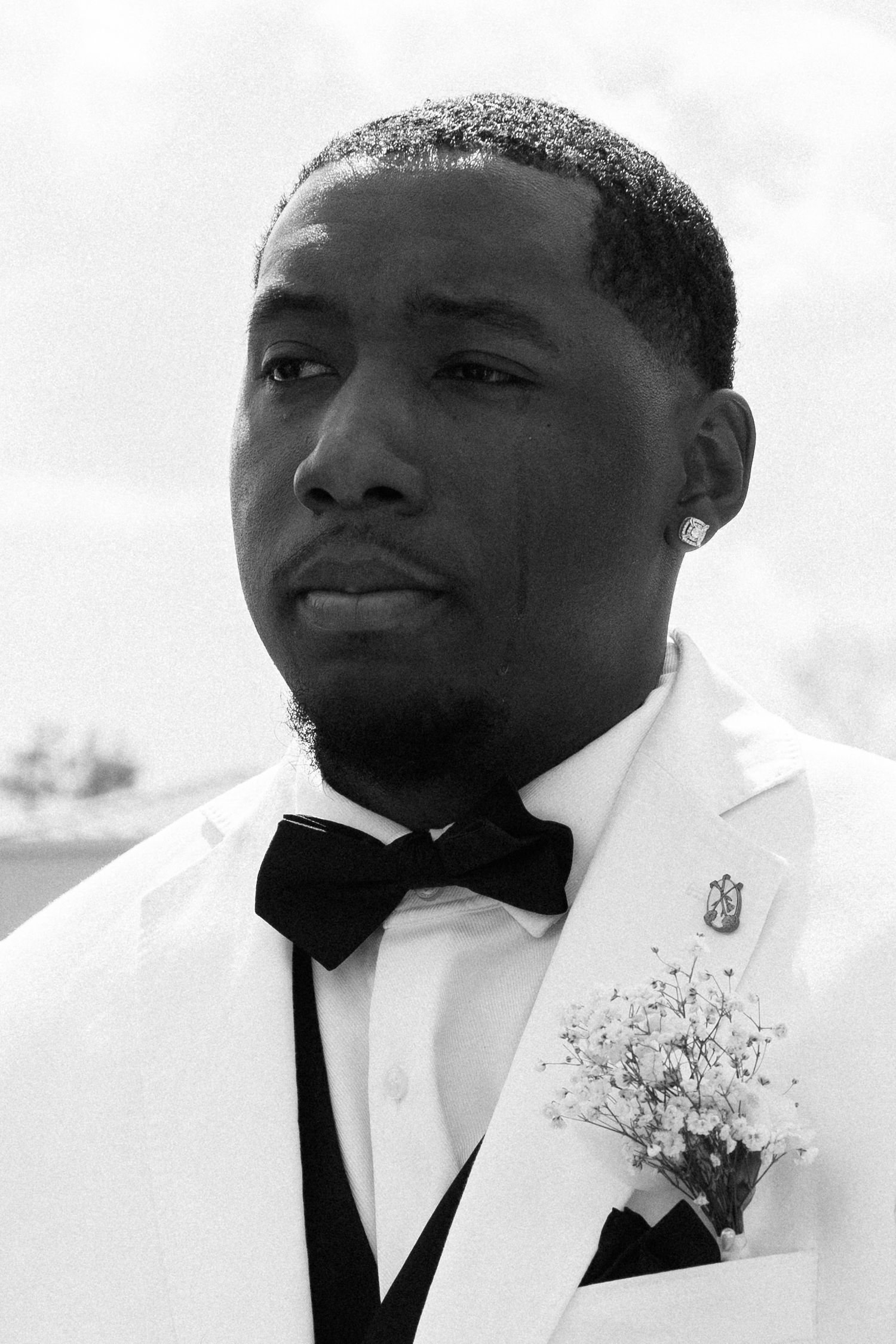 Black and white portrait of a groom in a white tuxedo jacket with black bow tie and baby's breath boutonniere.