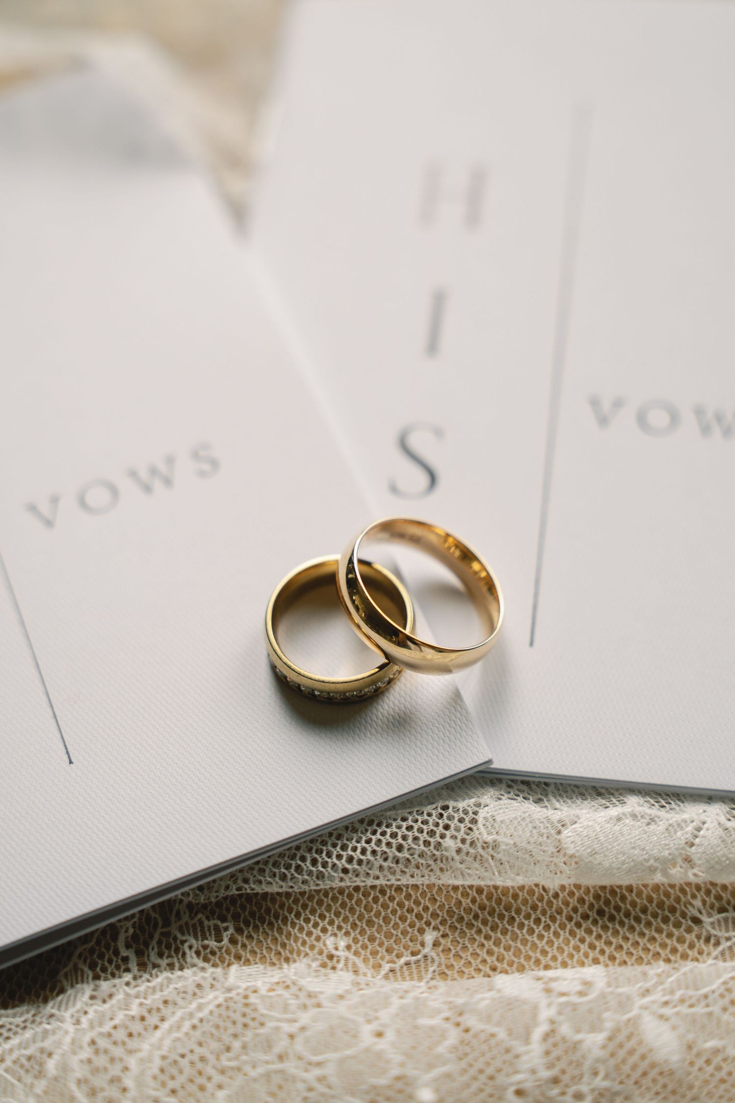 Two gold wedding bands rest on a white paper with the word 'vows' visible and delicate lace underneath.