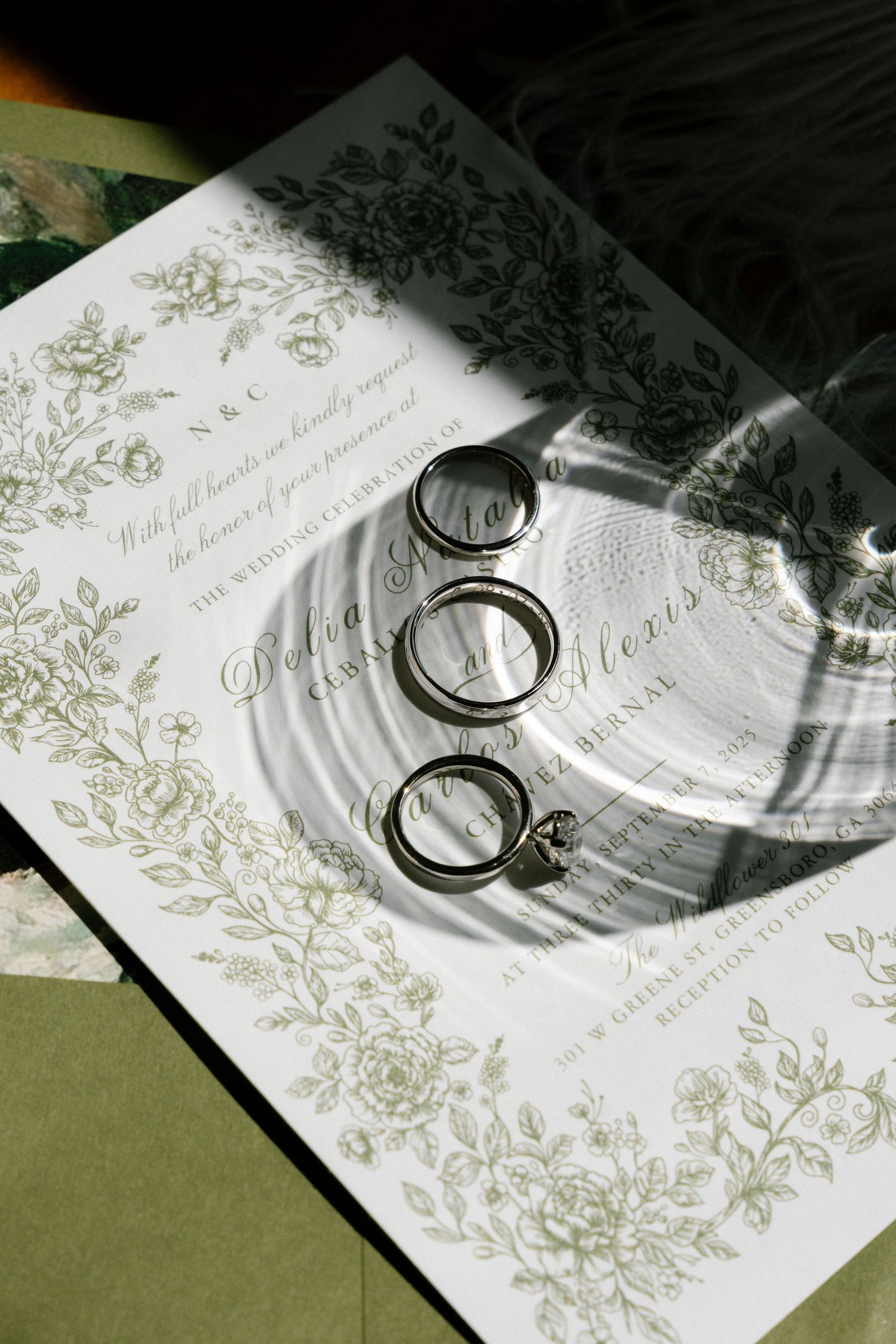 Wedding rings casting shadows on an elegant white invitation card with delicate green floral designs.