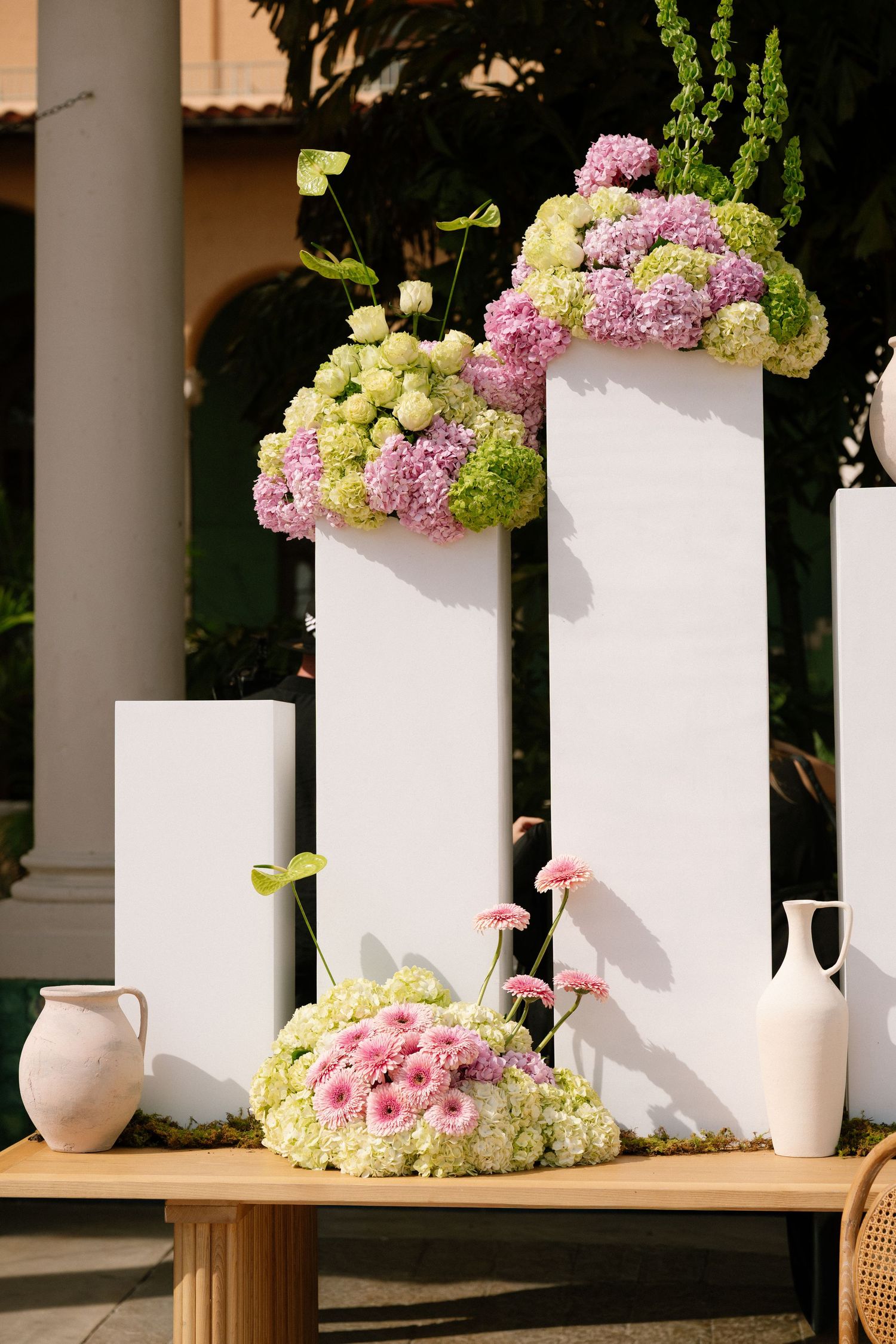 White pedestal display with pink and green hydrangea floral arrangements and ceramic vases at an outdoor wedding.