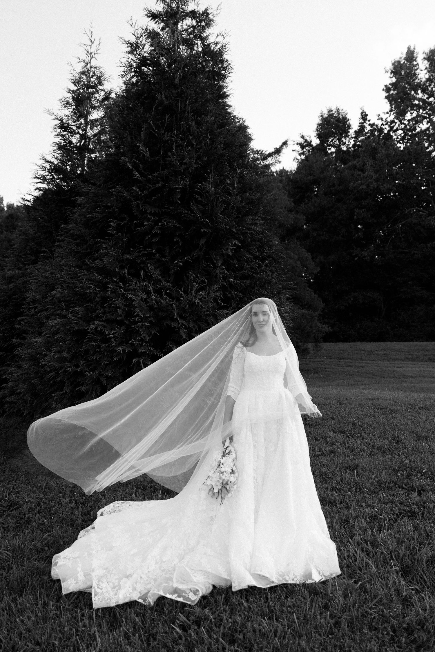 A flowing white wedding dress with a long veil billows gracefully across a grassy field with evergreen trees in the background.