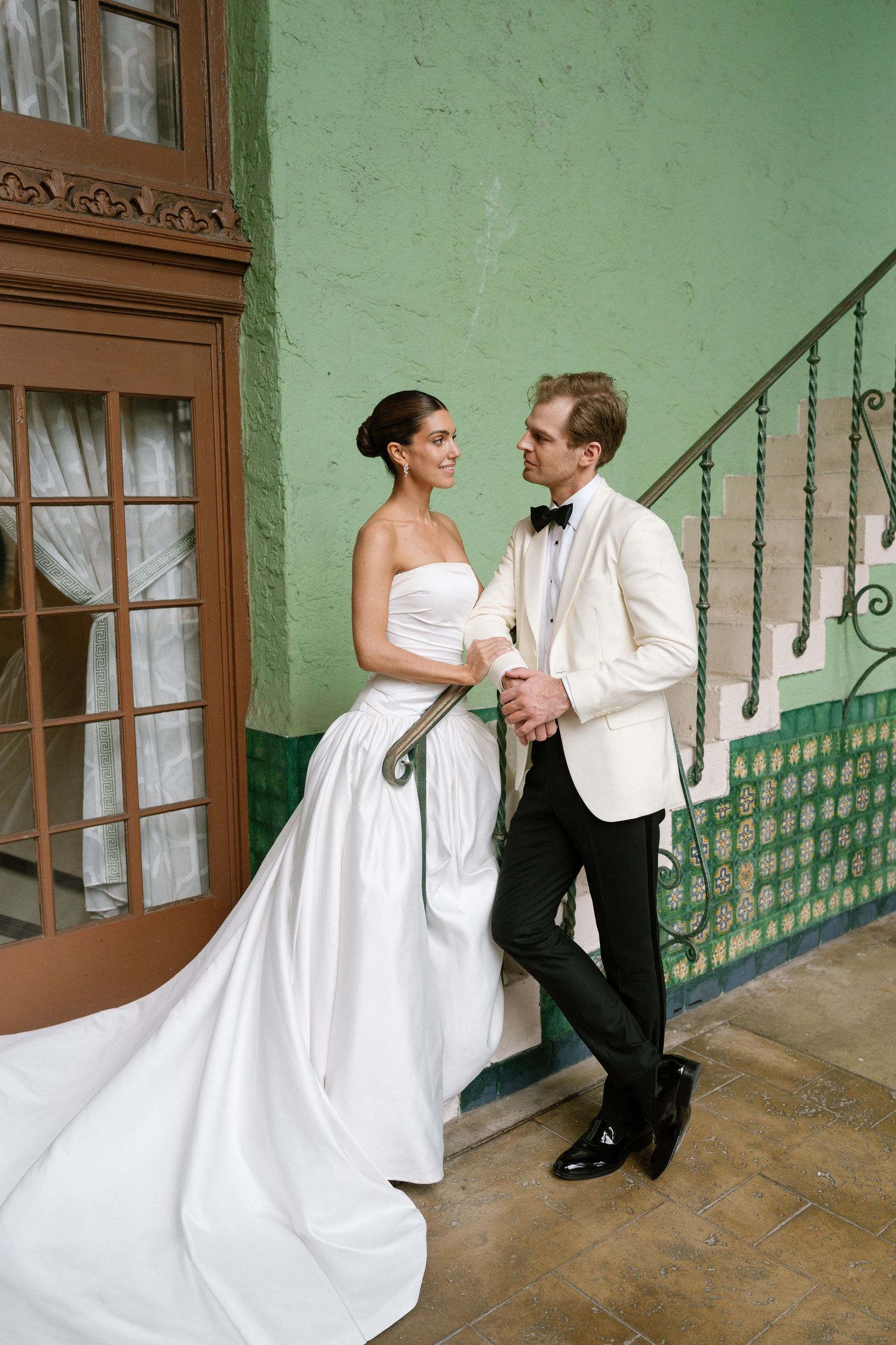 Bride and groom share romantic moment by ornate staircase, green tiled walls, bride in flowing white gown.